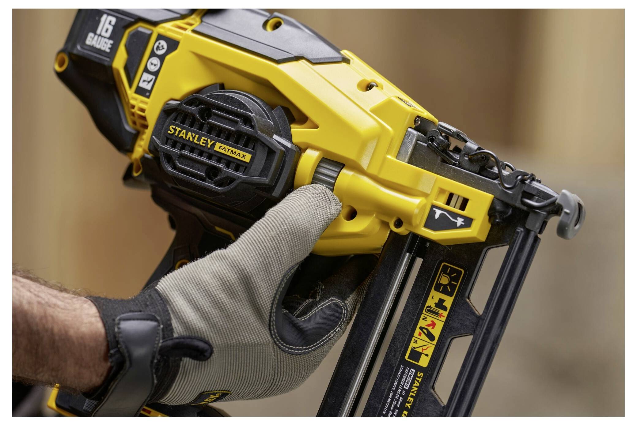 Eine Person mit Handschuh hält einen gelben 16er-Druckluftnagler in der Hand und bereitet ihn zur Verwendung vor. Sicherheitssymbole und der Markenname sind auf dem Werkzeug sichtbar.