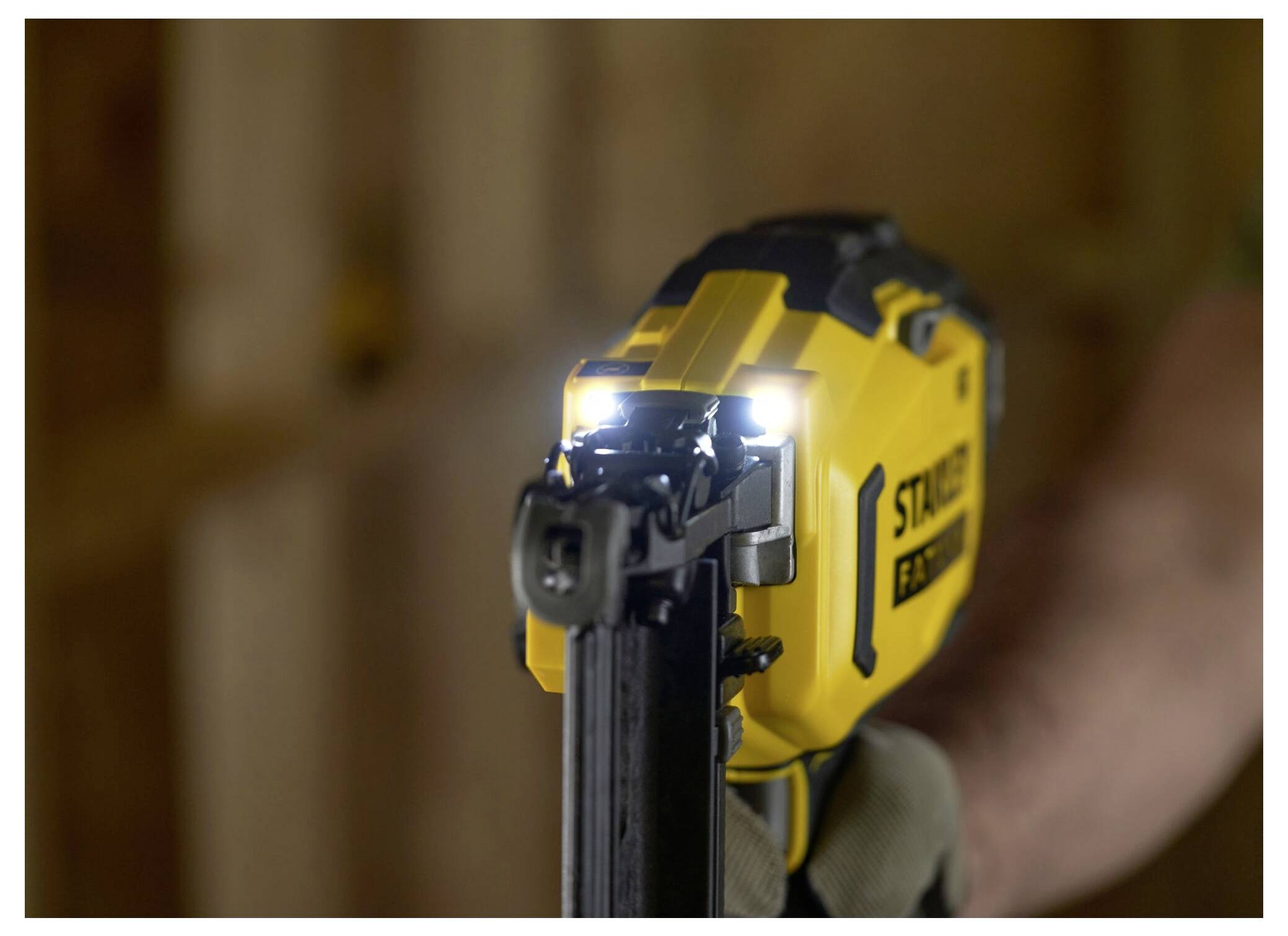 Eine Nahaufnahme einer Arbeiterhand, die eine gelbe Nagelschussmaschine mit LED-Leuchten hält, fokussiert auf ihre Düse, in einer schwach beleuchteten Baustelle.