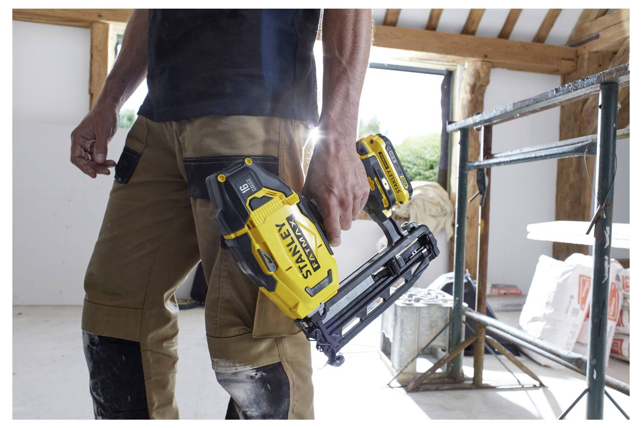 Eine Person in einer Werkstatt hält eine gelb-schwarze Nagelspistole und trägt braune Arbeitshosen und Knieschützer. Im Hintergrund sind Gerüste und Werkzeuge sichtbar.