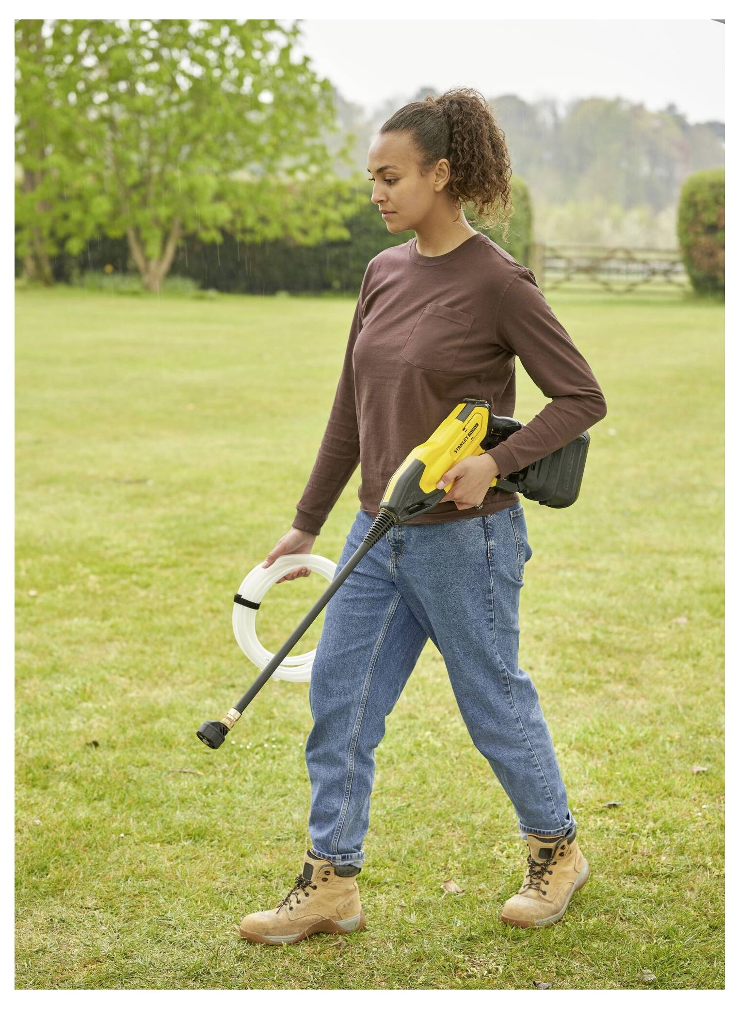 Eine Person in Freizeitkleidung geht über Gras und trägt einen gelben Hochdruckreiniger und einen Schlauch, mit Bäumen und einem Zaun im Hintergrund.