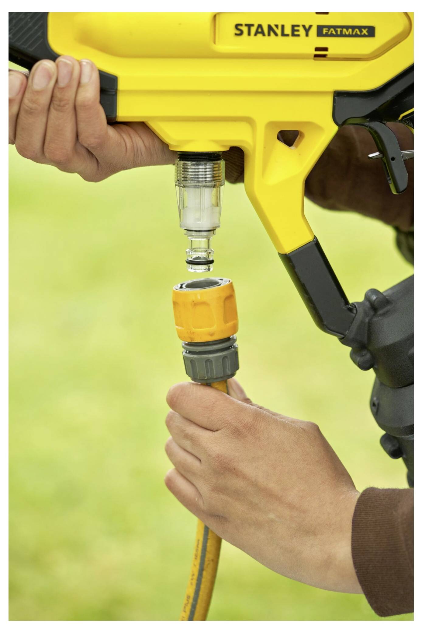Eine Person befestigt einen gelben Schlauch an einem gelben Stanley Fatmax Elektrowerkzeug, im Freien mit unscharfem grünem Hintergrund.