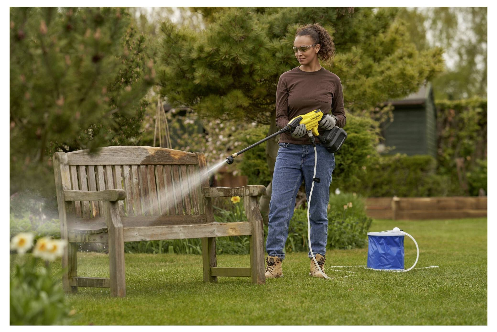 Eine Person, die Handschuhe und Schutzbrille trägt, benutzt einen Hochdruckreiniger, um eine Holzbank in einem Garten zu reinigen. Die Umgebung ist von verschiedenen Bäumen und Blumen umgeben.