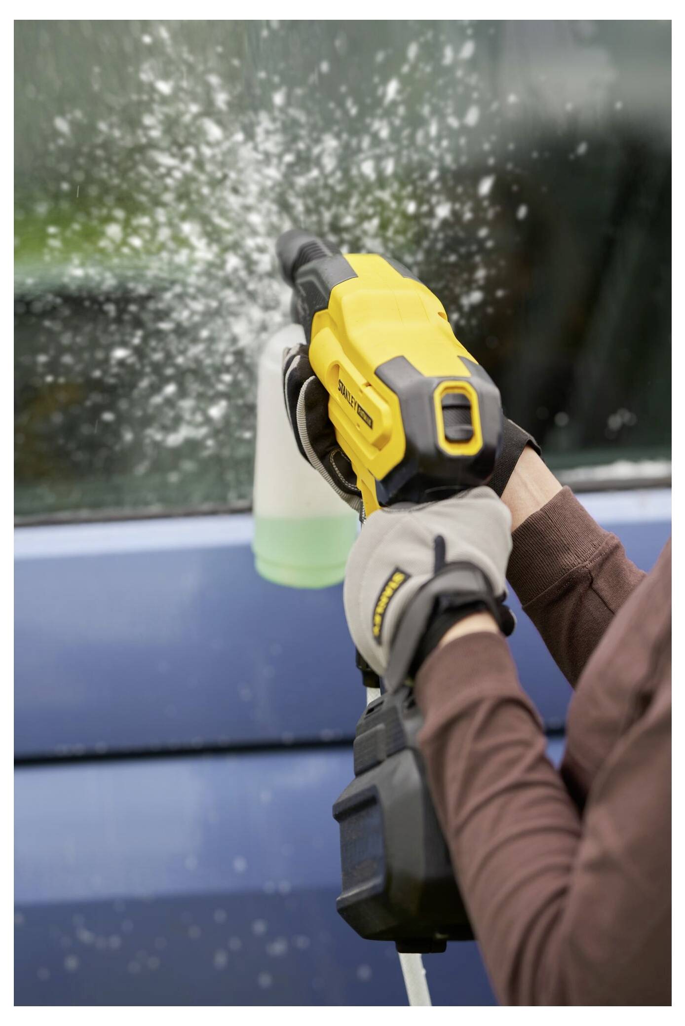 Eine Person benutzt einen gelben Hochdruckreiniger mit einer Seifenspenderanlage, um ein Auto zu reinigen, und trägt dabei Handschuhe zum Schutz.