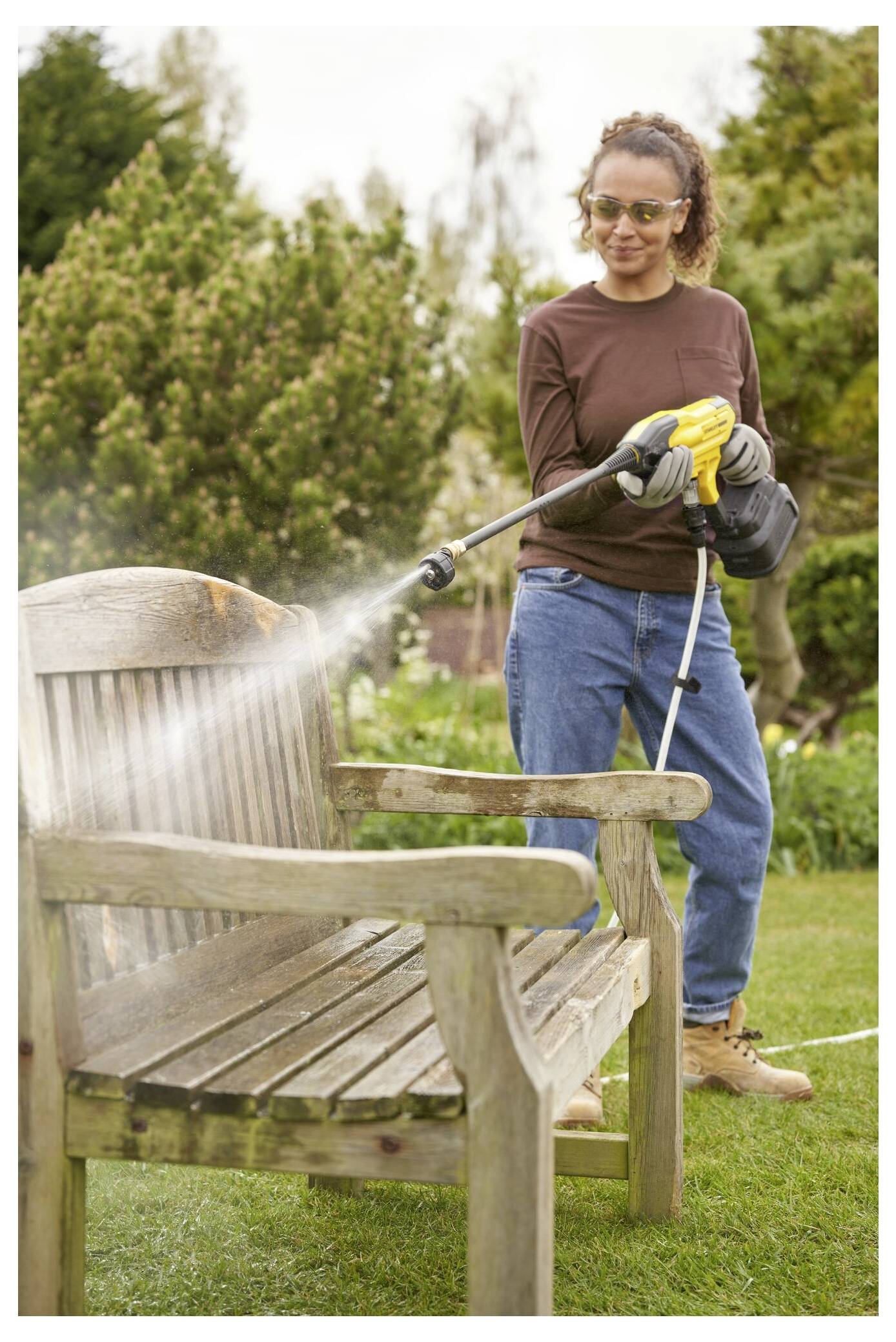 Eine Person mit Brille und Handschuhen nutzt einen Hochdruckreiniger, um eine Holzbank in einer Gartenumgebung mit Bäumen im Hintergrund zu reinigen.