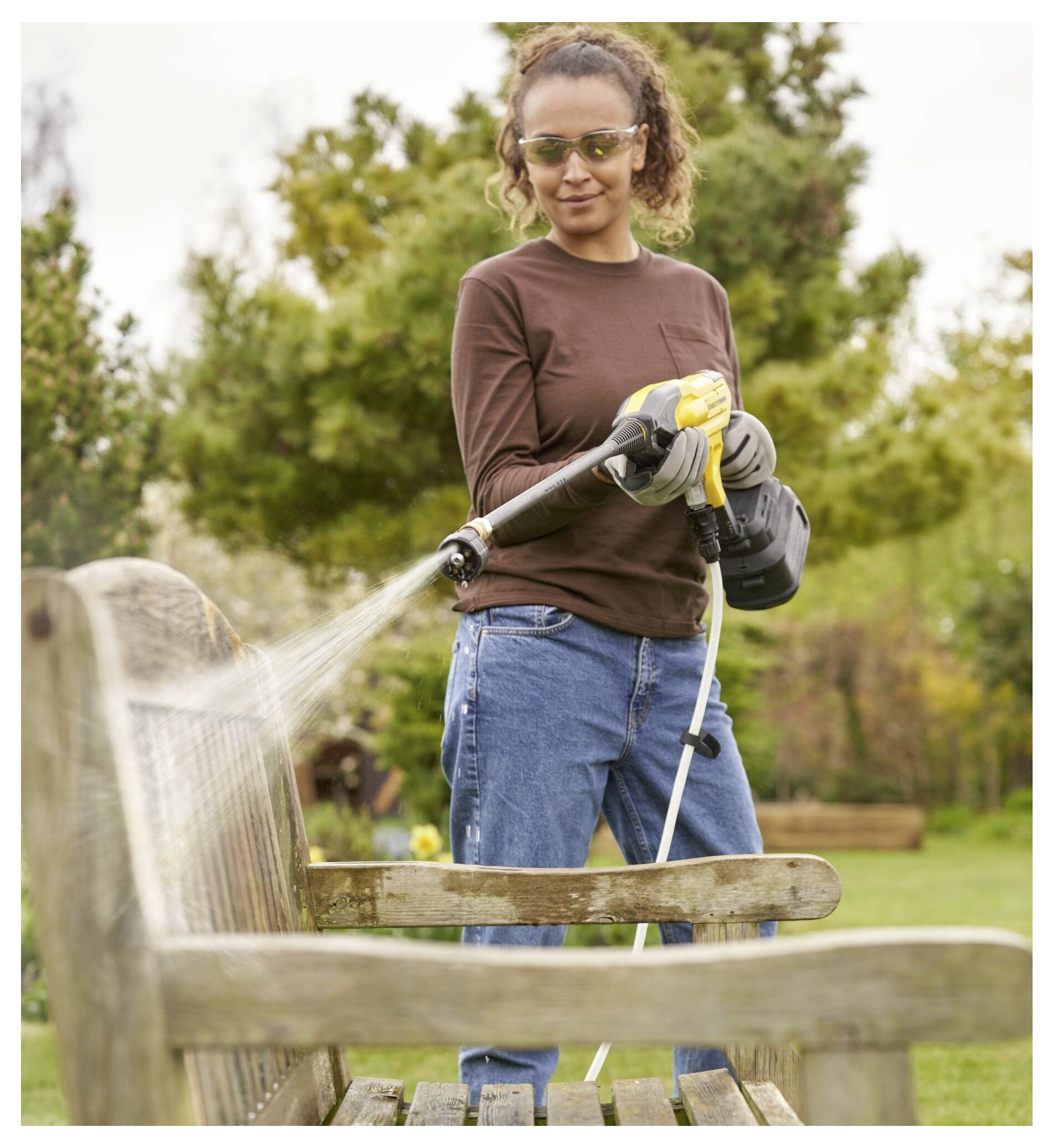 Eine Person in Schutzbrille und Handschuhen reinigt eine hölzerne Gartenbank mit einem Hochdruckreiniger in einer Gartenumgebung.