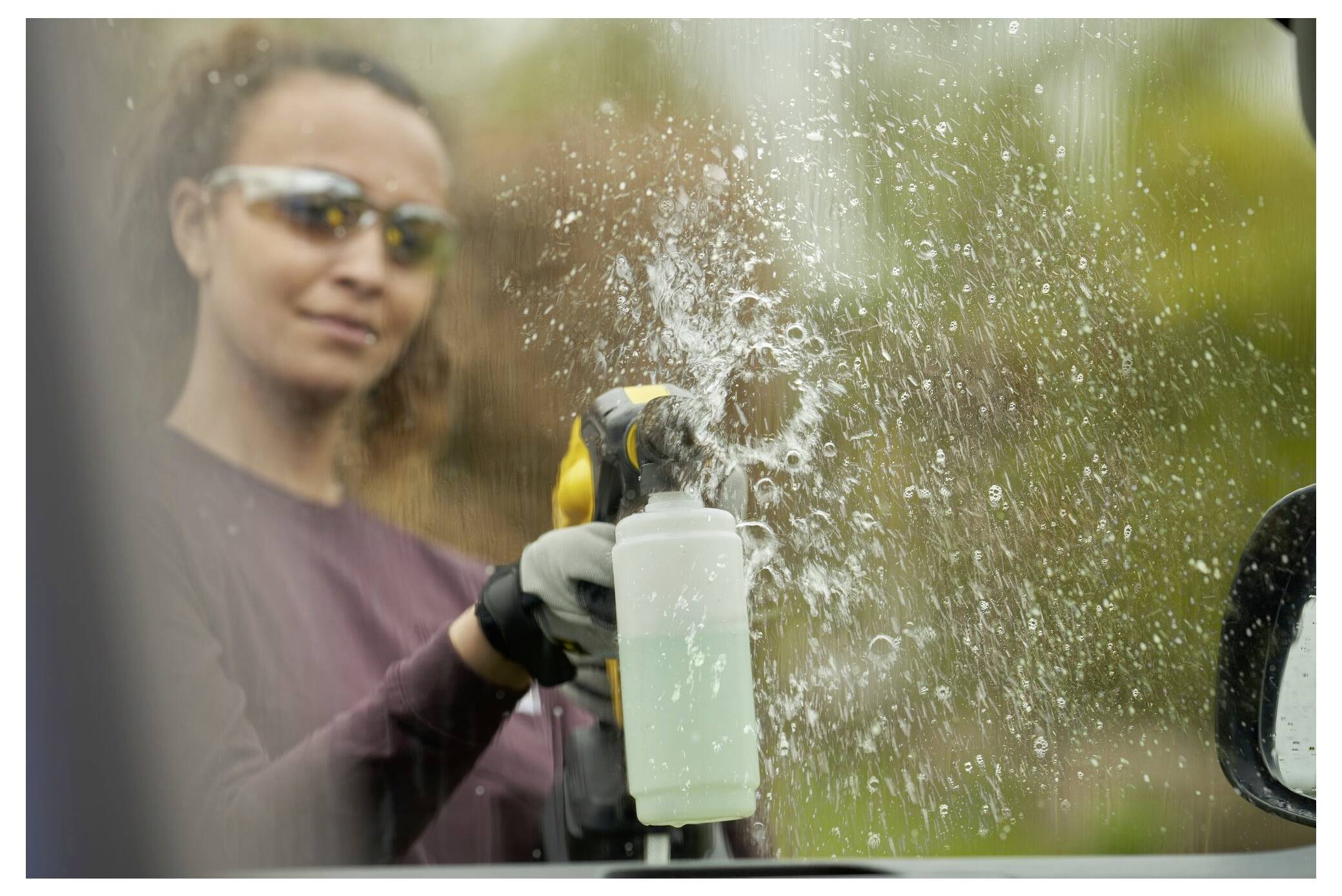 Eine Person, die eine Schutzsonnenbrille trägt, reinigt ein Fenster mit einer Sprühflasche, wobei sichtbare Wassertropfen auf der Glasoberfläche entstehen.