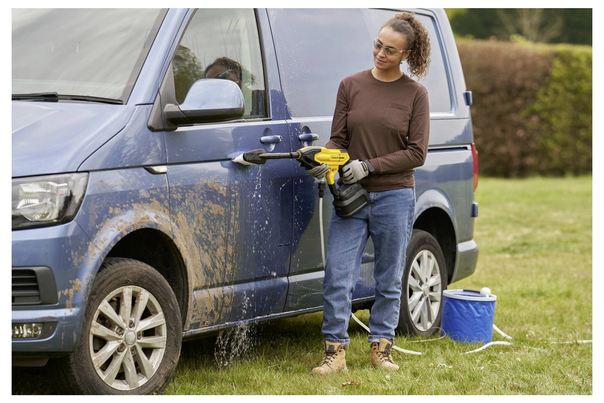Eine Person wäscht einen schmutzigen blauen Lieferwagen mit einem tragbaren Hochdruckreiniger auf Gras neben einem großen Eimer.