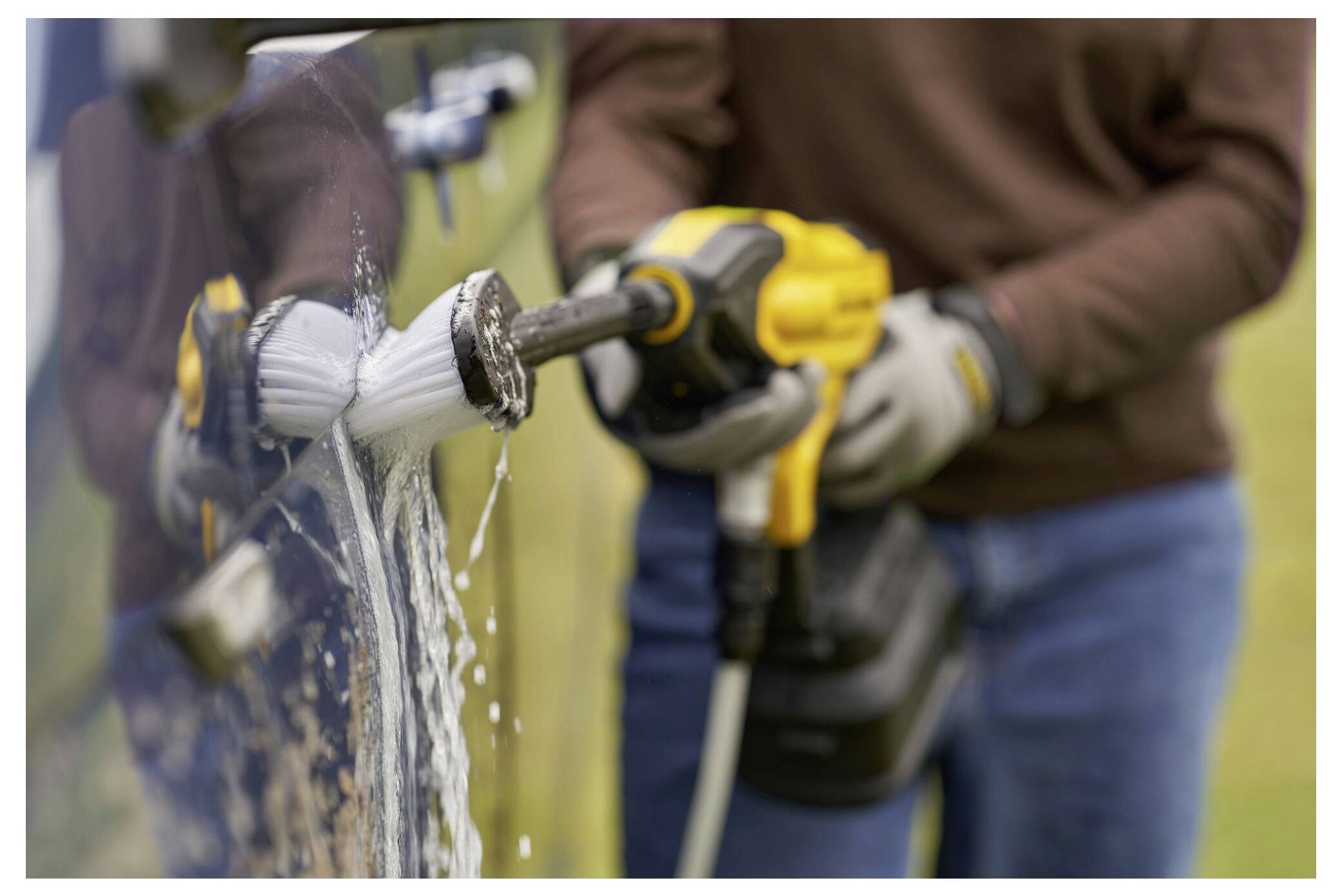 Eine Person verwendet einen Elektroschrauber mit einer Bürstenaufsatzvorrichtung, um die Seite eines Fahrzeugs zu reinigen, wobei Wasser und Seife sichtbar auf der Oberfläche wirken.