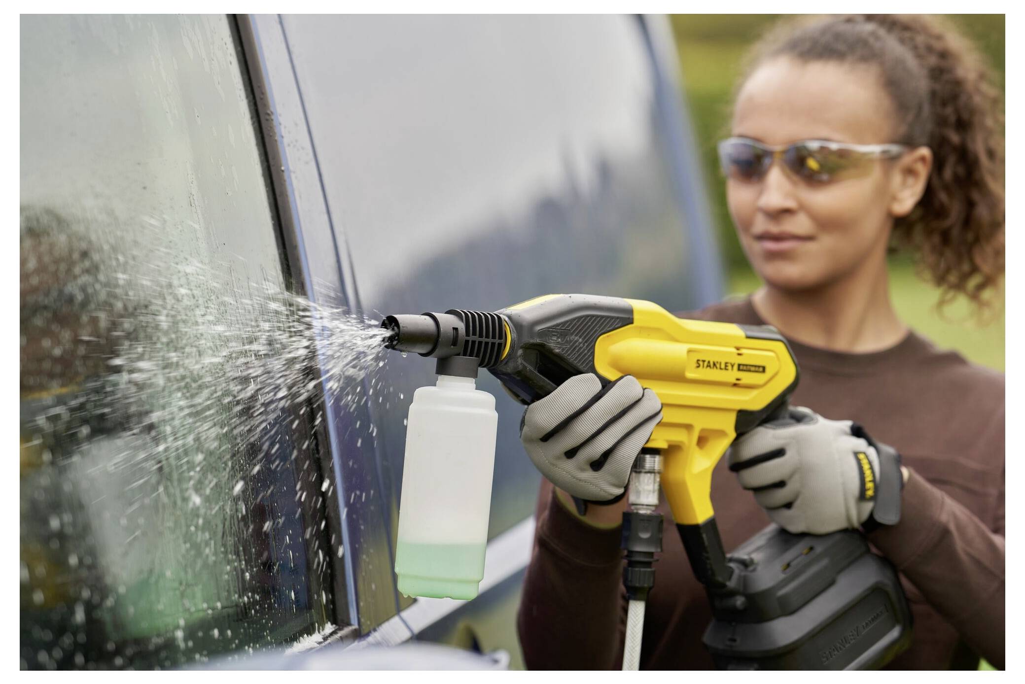 Eine Person nutzt einen gelben Hochdruckreiniger, um das Fenster eines Fahrzeugs im Freien zu reinigen, während sie Schutzhandschuhe und eine Schutzbrille trägt.