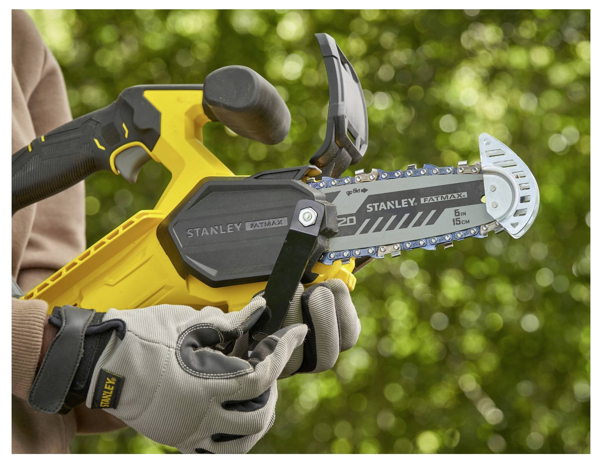 Eine Person mit Handschuhen hält eine gelb-schwarze handliche Kettensäge mit sichtbaren Details auf dem Blatt, im Freien vor einem grünen, unscharfen Hintergrund.