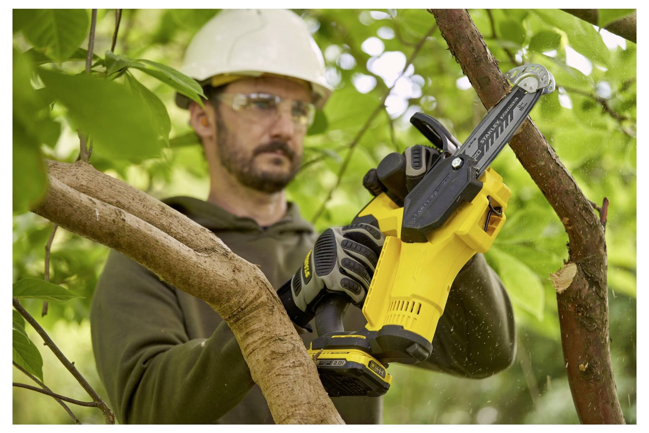 Eine Person, die Schutzausrüstung trägt, benutzt eine gelbe Kettensäge, um einen Baumast in einer belaubten Außenumgebung zu schneiden.
