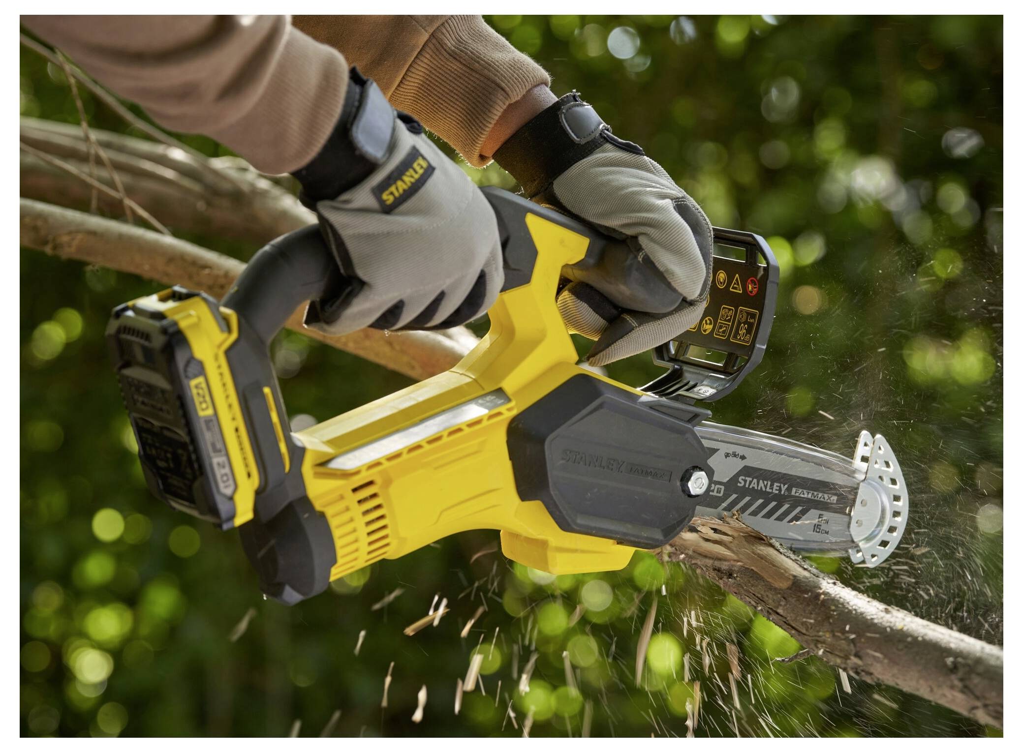 Eine Person benutzt eine gelbe kabellose Kettensäge, um einen Baumast im Freien zu schneiden, wobei Sägespäne fliegen. Die Szene vermittelt eine Außenarbeitstätigkeit.