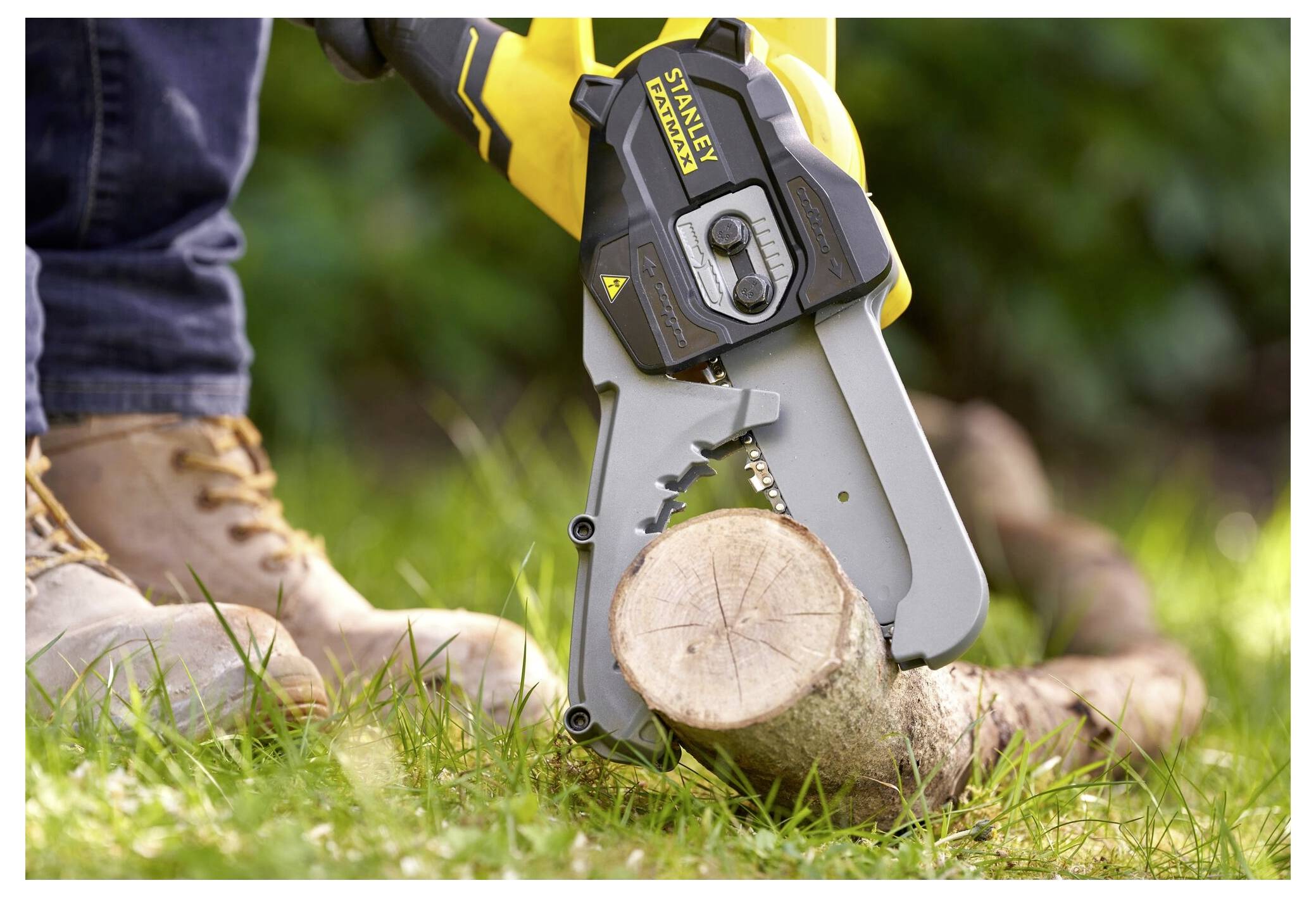 Eine Person in Arbeitsschuhen benutzt eine tragbare elektrische Kettensäge, um einen Baumstamm auf grasbewachsenem Boden zu schneiden. Das Gerät ist markengebunden und gelb.