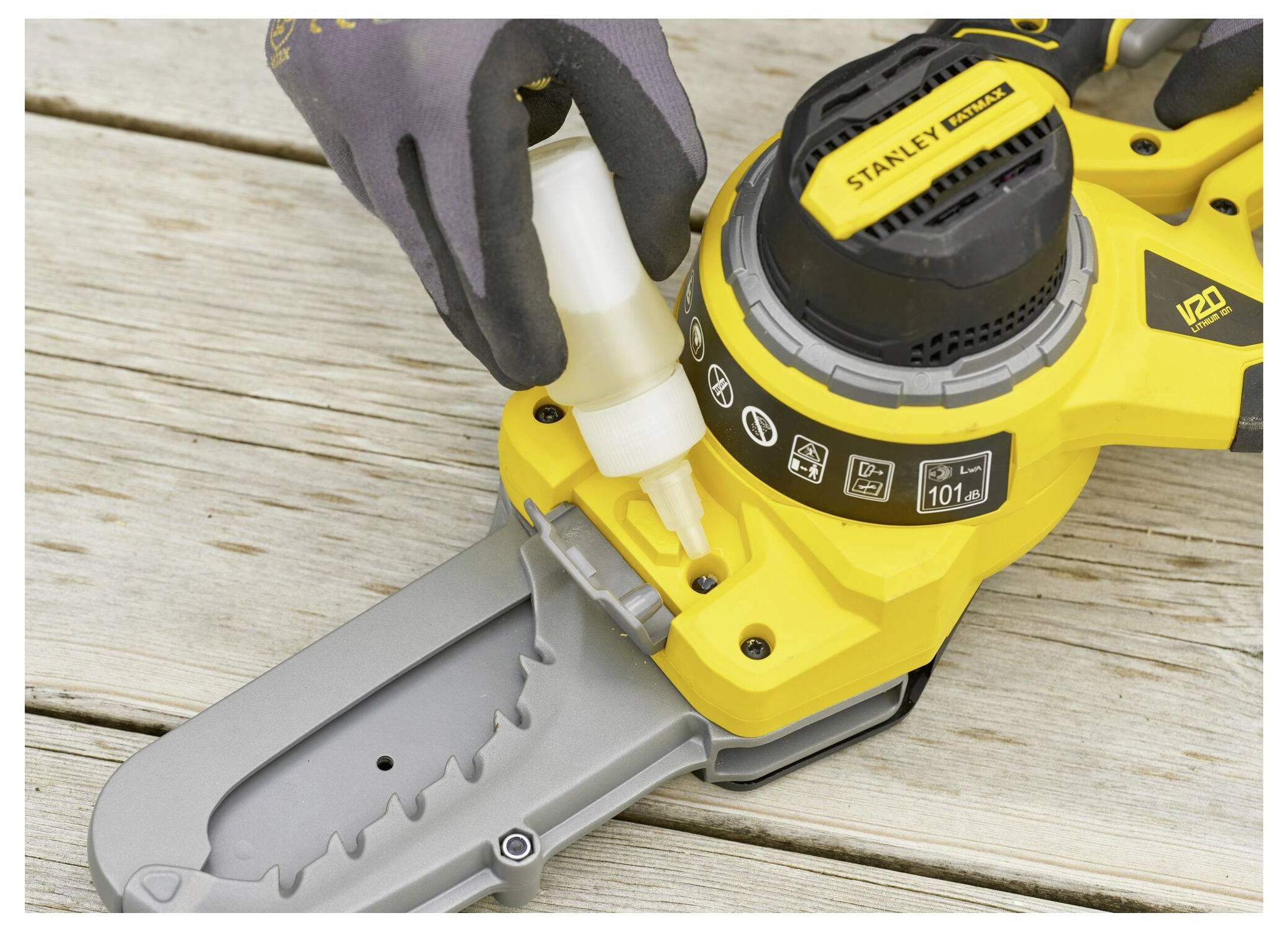 Eine Person gibt Öl in eine gelbe Kettensägeschiene. Die Säge ist auf einer Holzoberfläche positioniert. Handschuhe werden zum Schutz getragen.