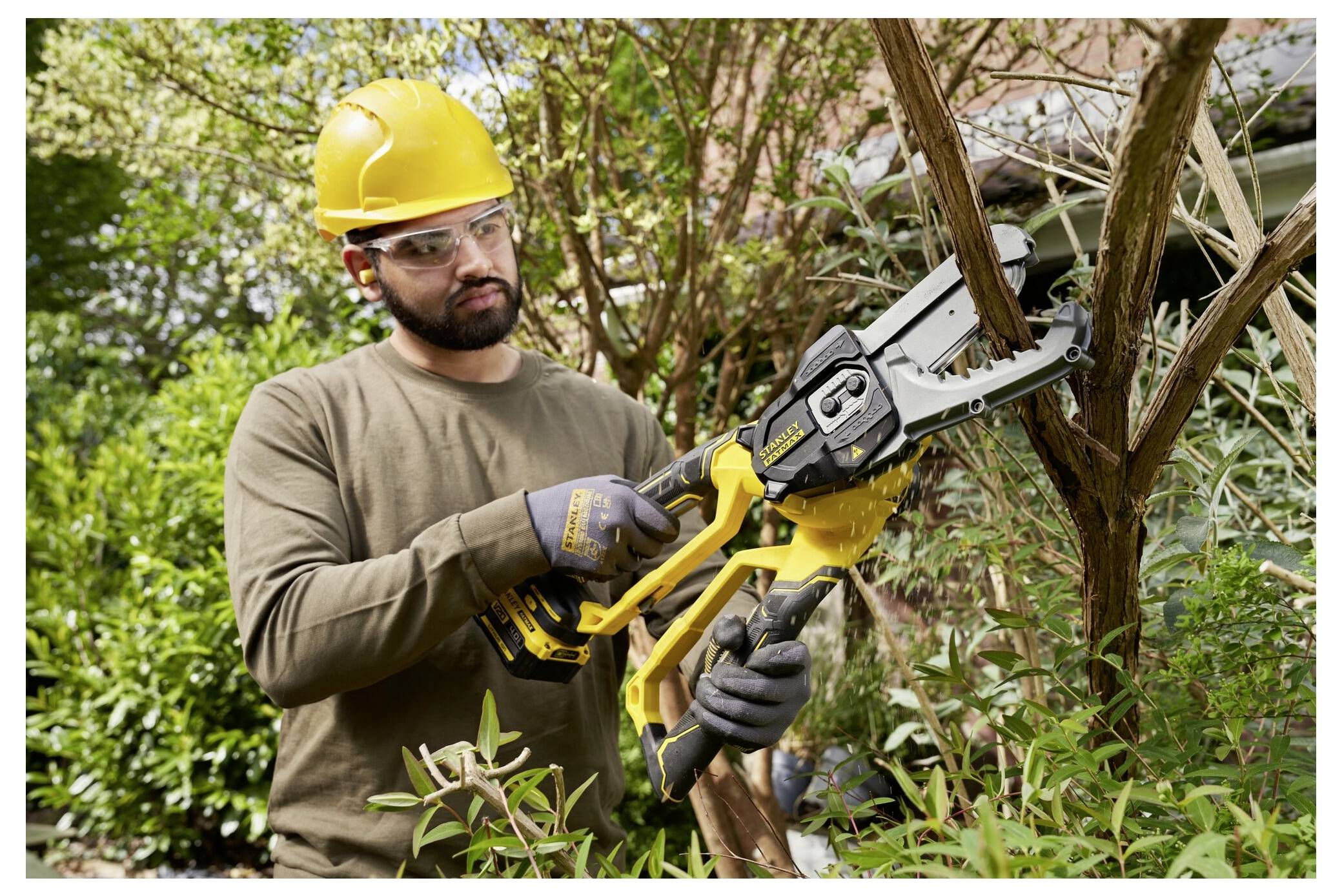 Eine Person, die einen gelben Schutzhelm und eine Schutzbrille trägt, benutzt ein gelbes Elektrowerkzeug, um Äste in einem Garten zu schneiden.