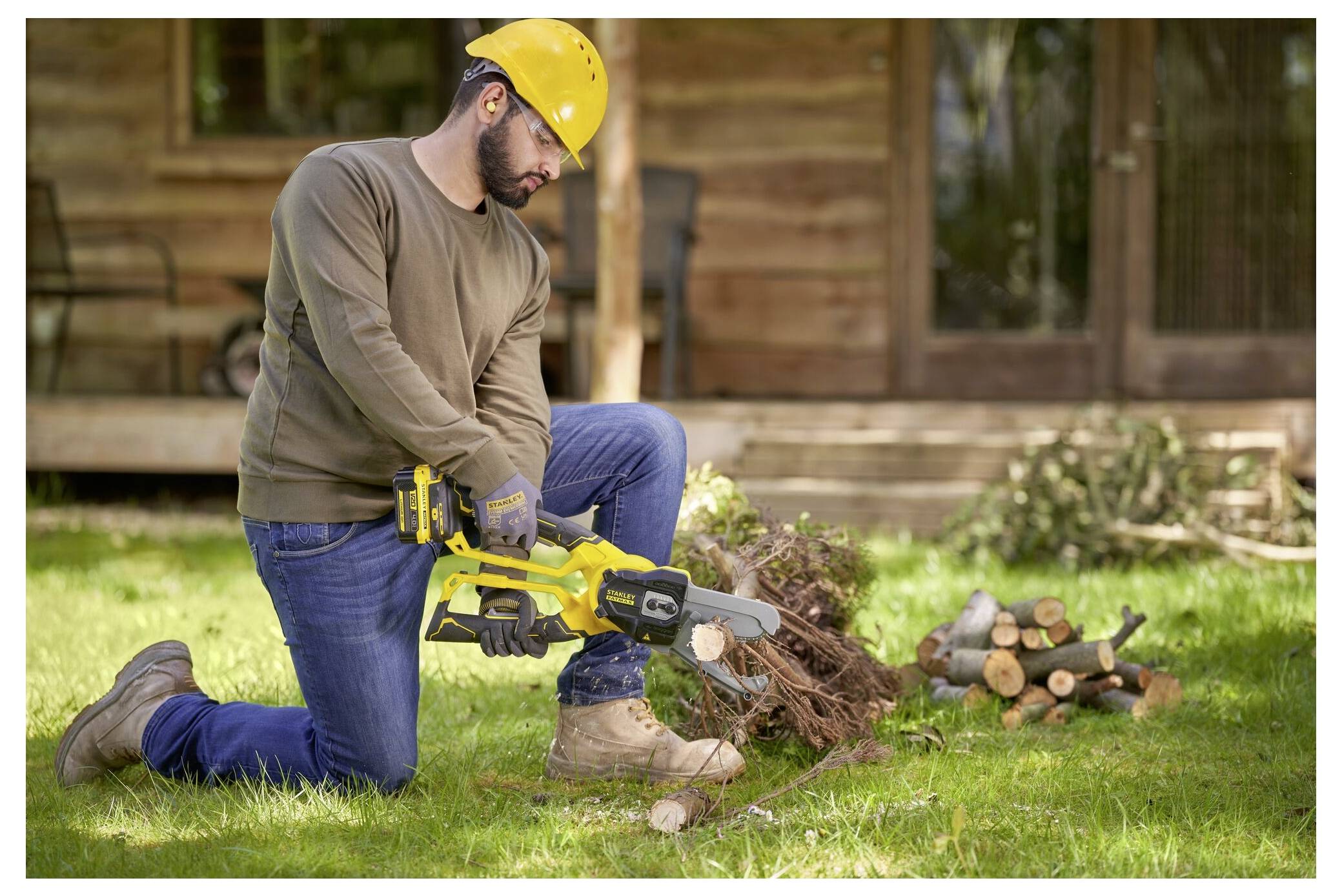 Eine Person trägt einen gelben Schutzhelm und benutzt eine Kettensäge, um einen großen Ast auf dem Boden zu schneiden, wobei ein Holzhaus im Hintergrund zu sehen ist.