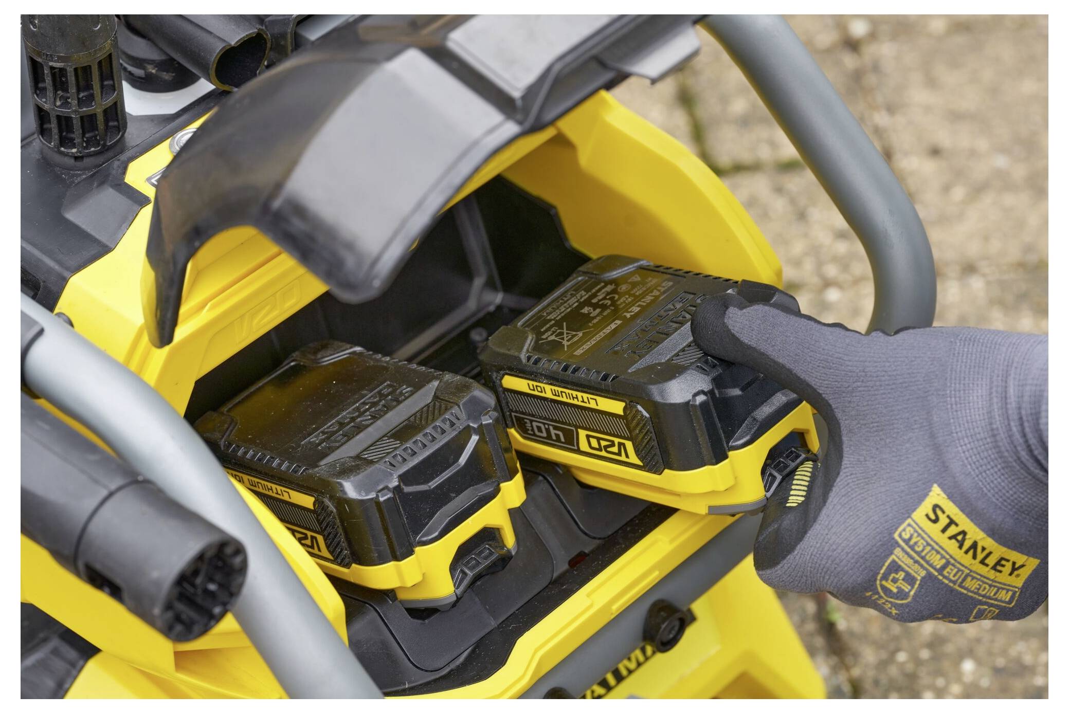 Die behandschuhte Hand einer Person führt eine Batterie in ein gelbes Stromwerkzeug-Ladegerät im Freien ein. Zwei Batteriefächer sind sichtbar, eines leer und eines belegt.