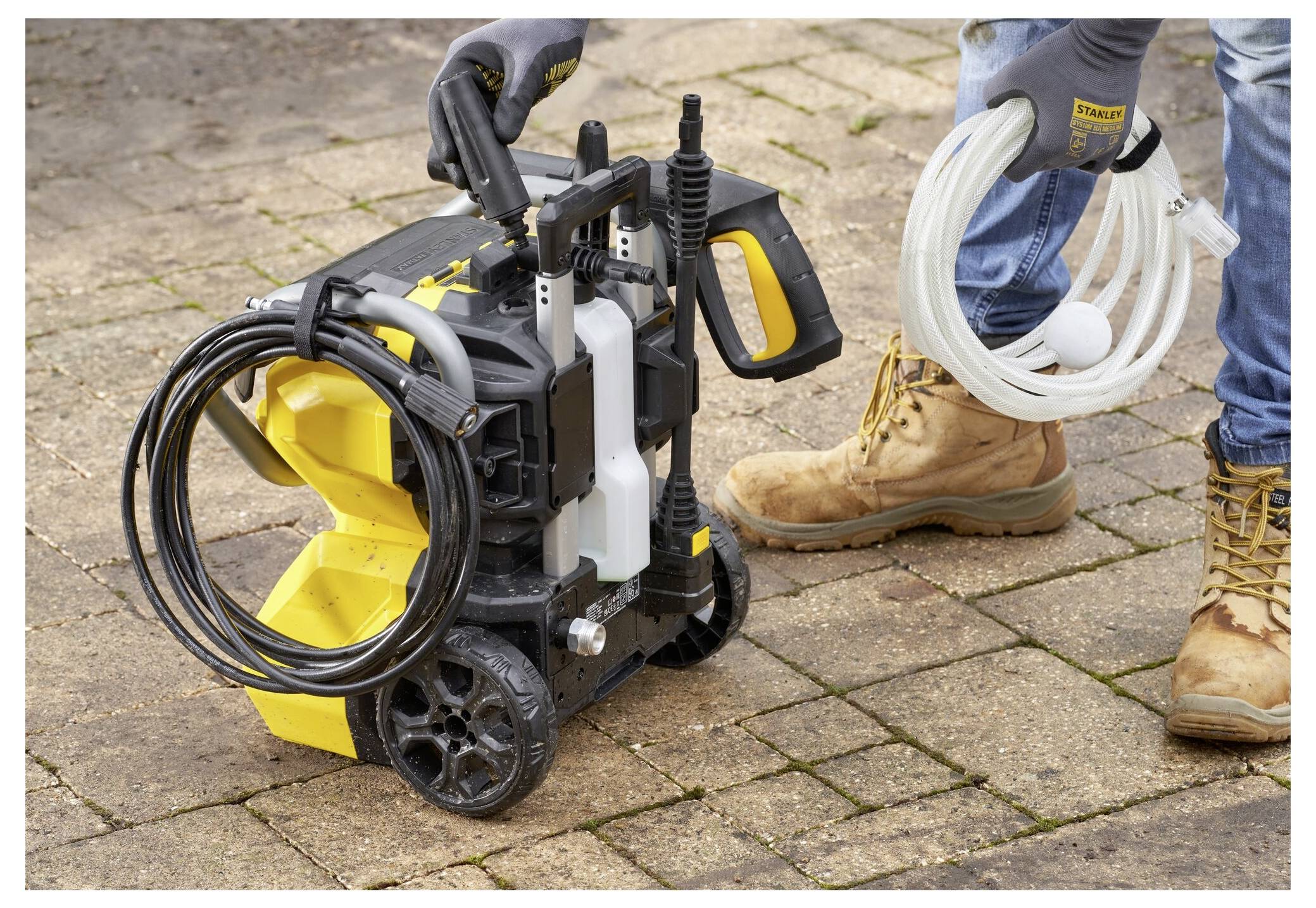 Eine Person mit Handschuhen hält einen tragbaren Hochdruckreiniger und einen Saugschlauch und steht auf einem Steinweg.
