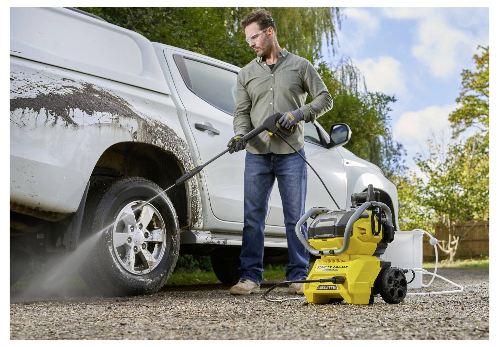 Eine Person trägt Handschuhe und Schutzbrille und nutzt einen gelben Hochdruckreiniger, um einen schlammigen Reifen eines weißen Fahrzeugs an einem sonnigen Tag zu reinigen.