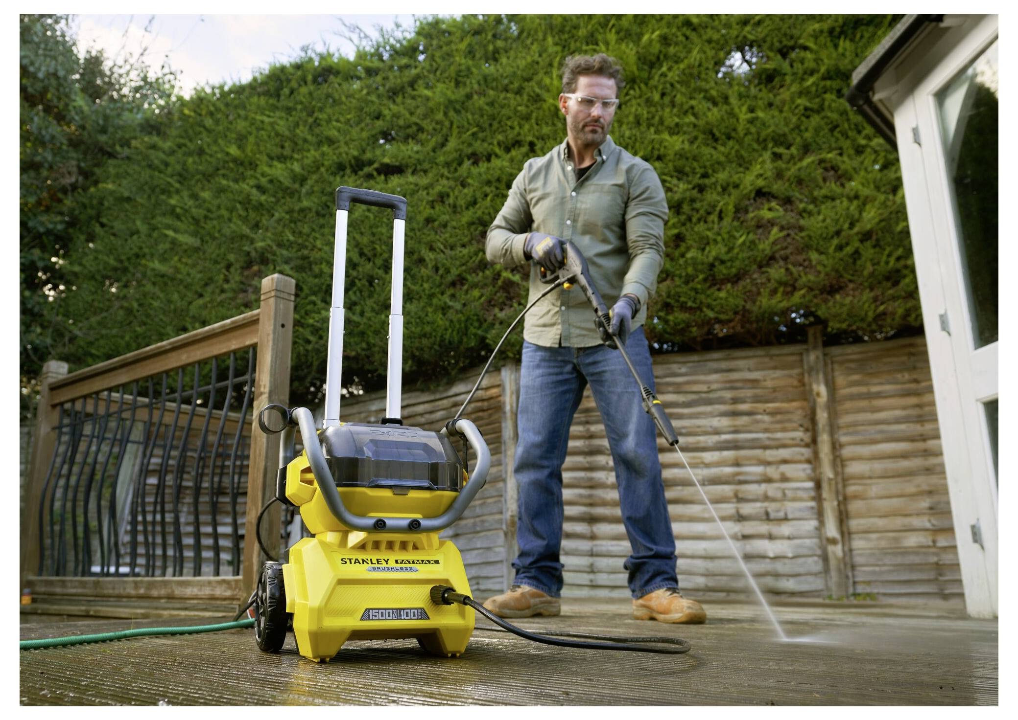 Ein Mann benutzt einen gelben Hochdruckreiniger, um ein Holzdeck im Freien zu reinigen, das von einem Holzzaun und Grünflächen im Hintergrund umgeben ist.