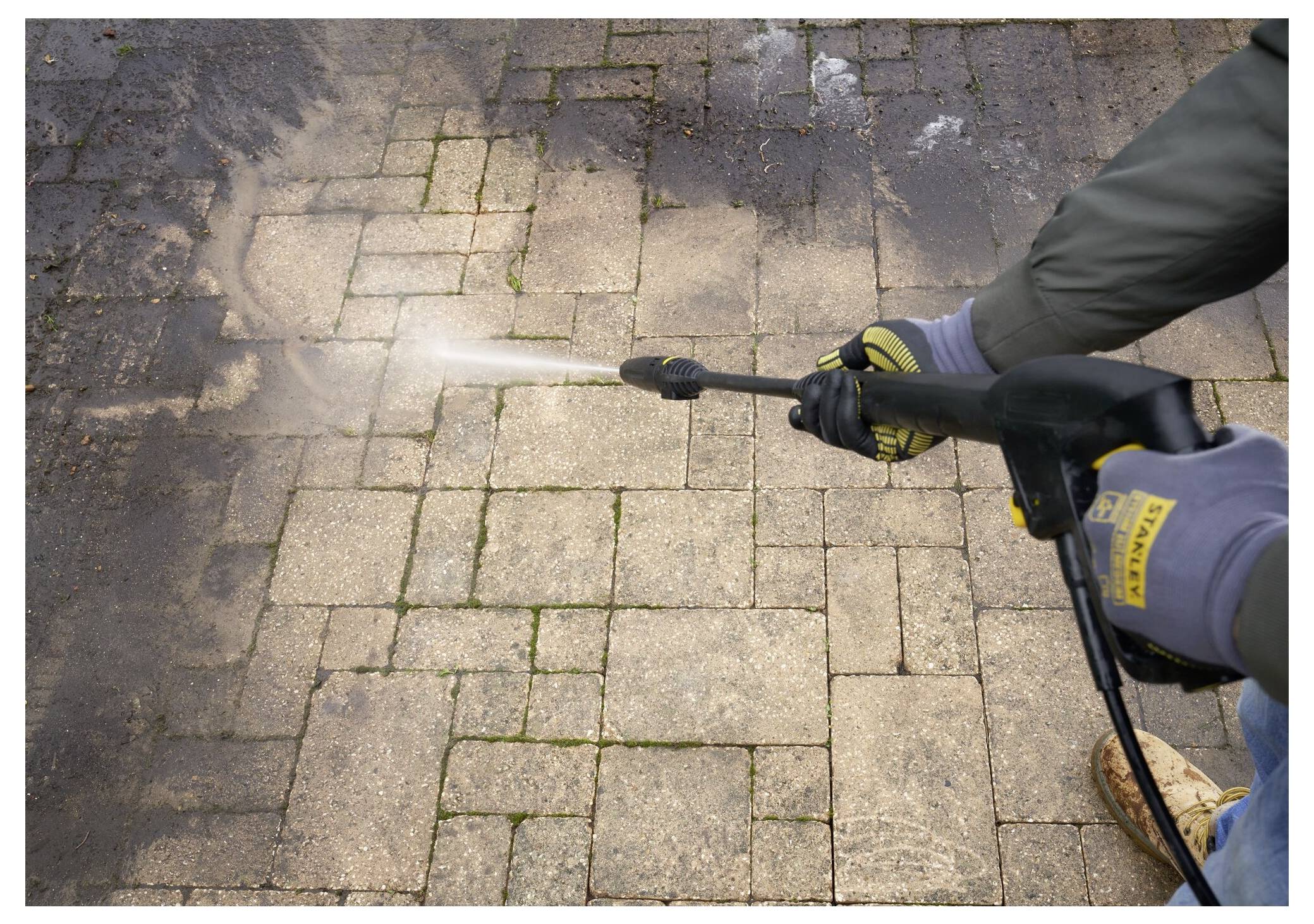 Eine Person benutzt einen Hochdruckreiniger, um eine Steinpflaster-Terrasse zu reinigen. Der Wasserstrahl entfernt Schmutz und offenbart darunter saubere Steine.
