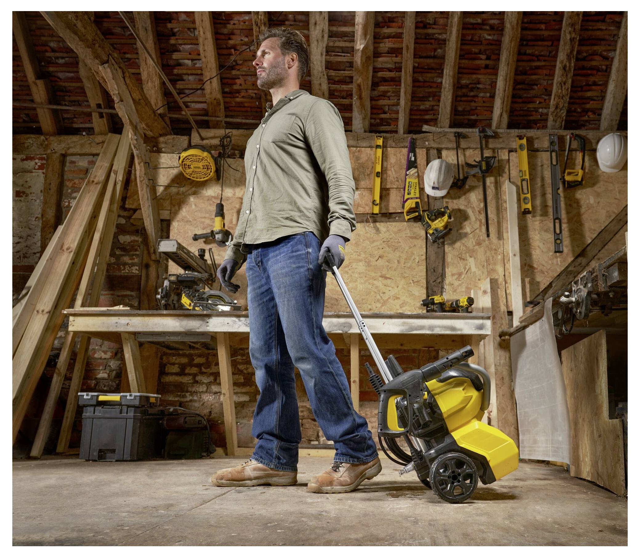 Eine Person in einer Werkstatt steht und hält ein gelbes Werkzeug auf Rädern, umgeben von verschiedenen Werkzeugen und Holzbalken, was auf aktive Arbeit hindeutet.