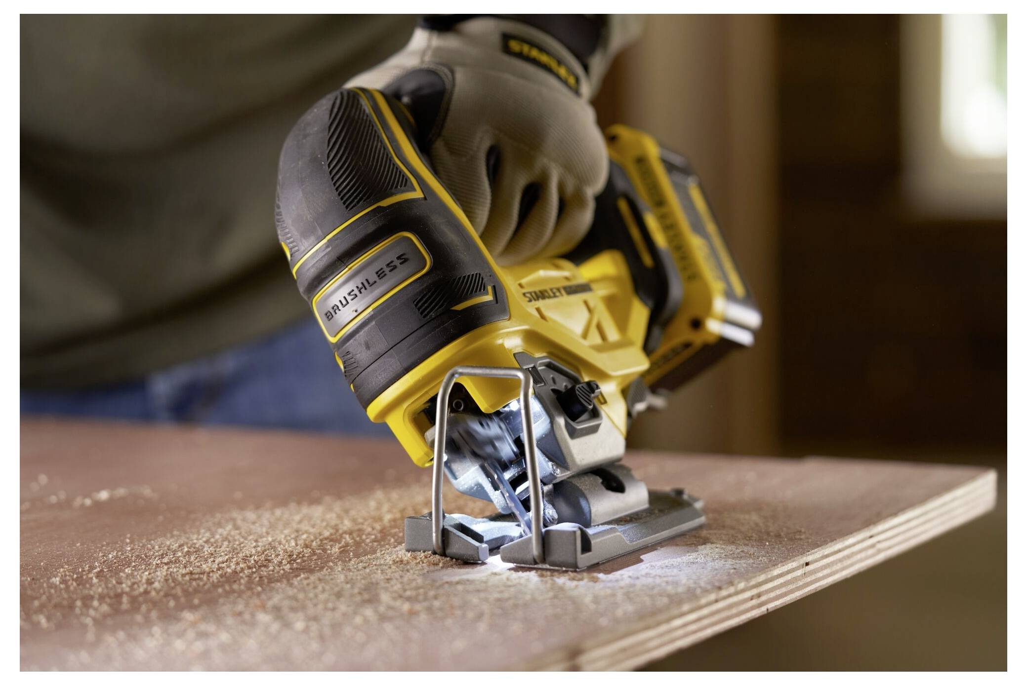 Eine Person benutzt eine gelbe kabellose Stichsäge, um ein Stück Sperrholz durchzuschneiden und dabei Sägespäne zu erzeugen. Der Fokus liegt auf dem Werkzeug und dem Prozess.