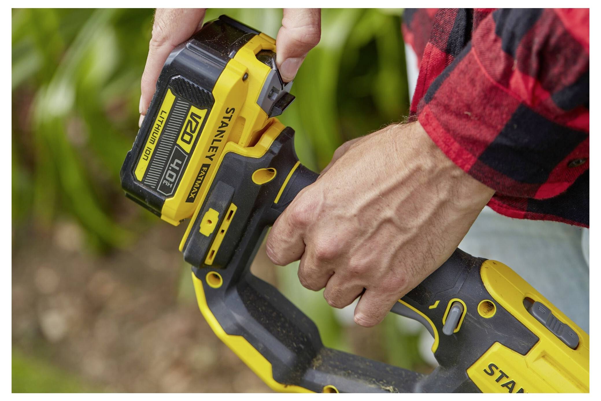 Eine Person in einem roten Karohemd setzt eine Lithium-Batterie in einen gelb-schwarzen Stanley-Akkubohrer ein, mit unscharfem grünem Hintergrund.