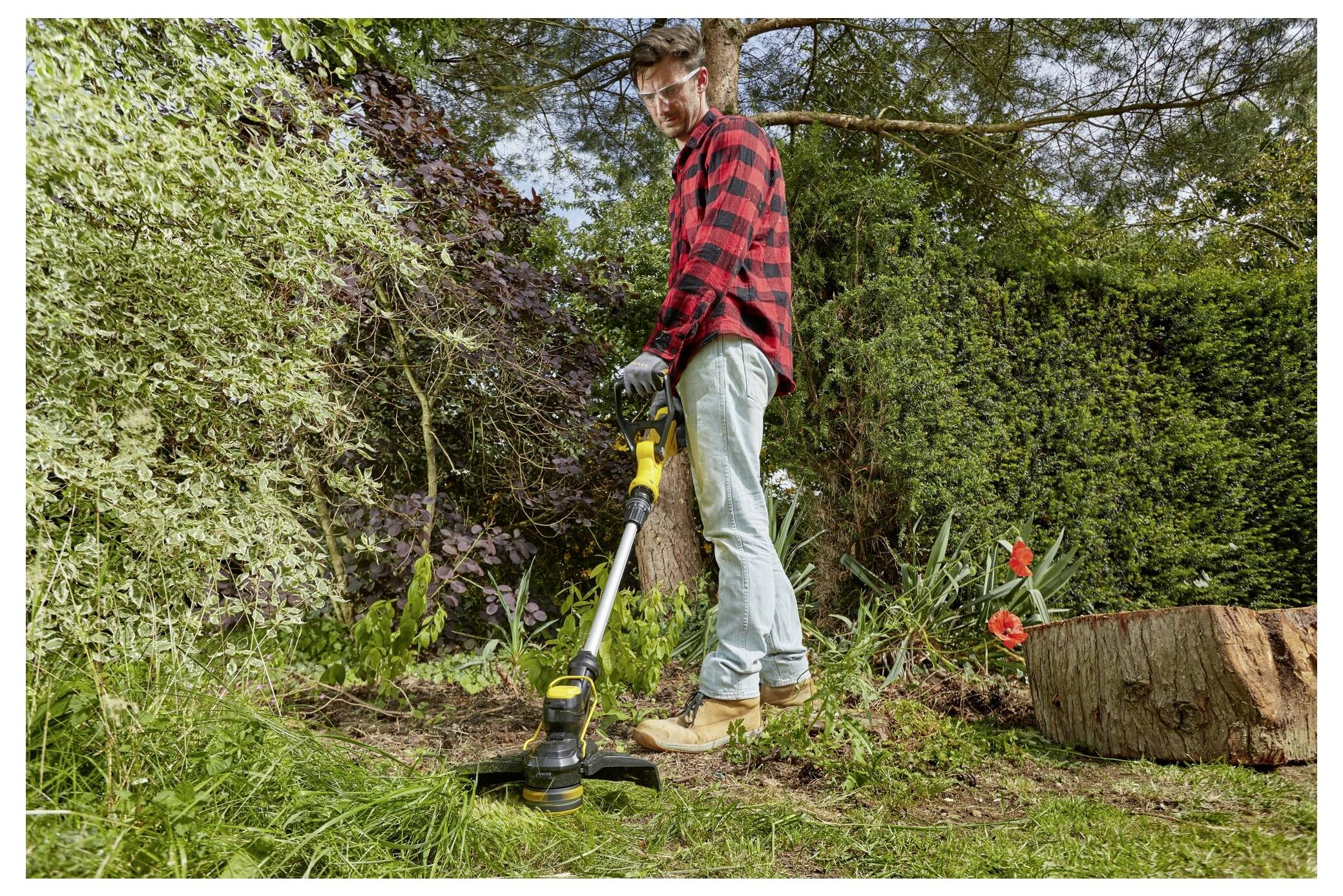 Ein Mann in einem roten karierten Hemd benutzt einen elektrischen Trimmer, um Gras in einem Garten zu schneiden, umgeben von Bäumen und Sträuchern.