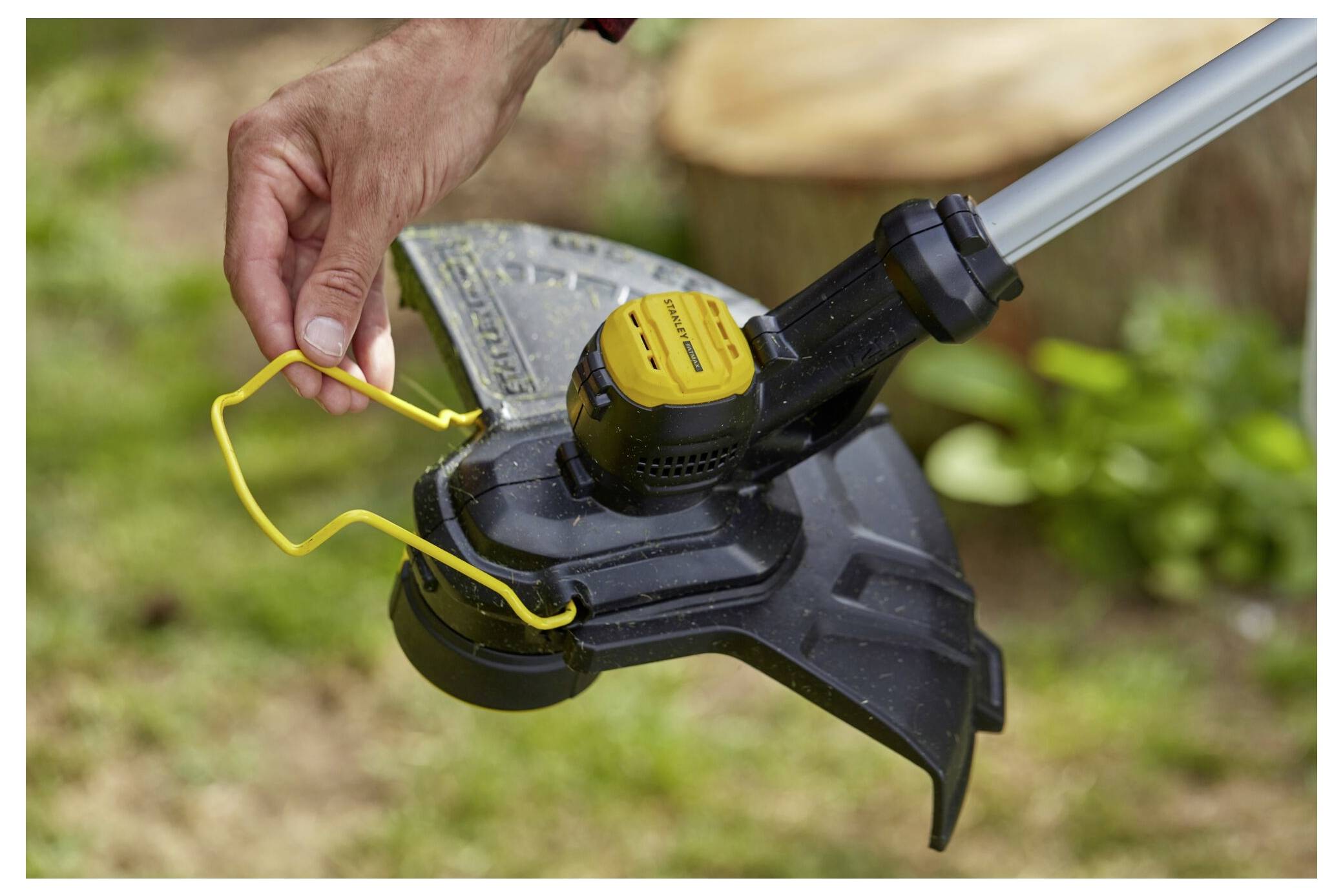 Eine Person hält den Kopf eines Fadentrimmer und stellt seine gelbe Schnur ein. Der Trimmer ruht auf grünem Gras mit einem Baumstumpf in der Nähe.