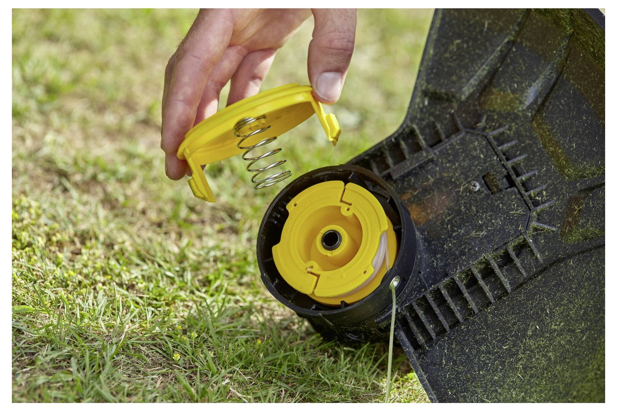 Eine Hand ersetzt eine gelbe Abdeckung und eine Feder auf einer Fadenspeule eines Rasentrimmers in einem Garten und zeigt die Wartung von Gartengeräten.