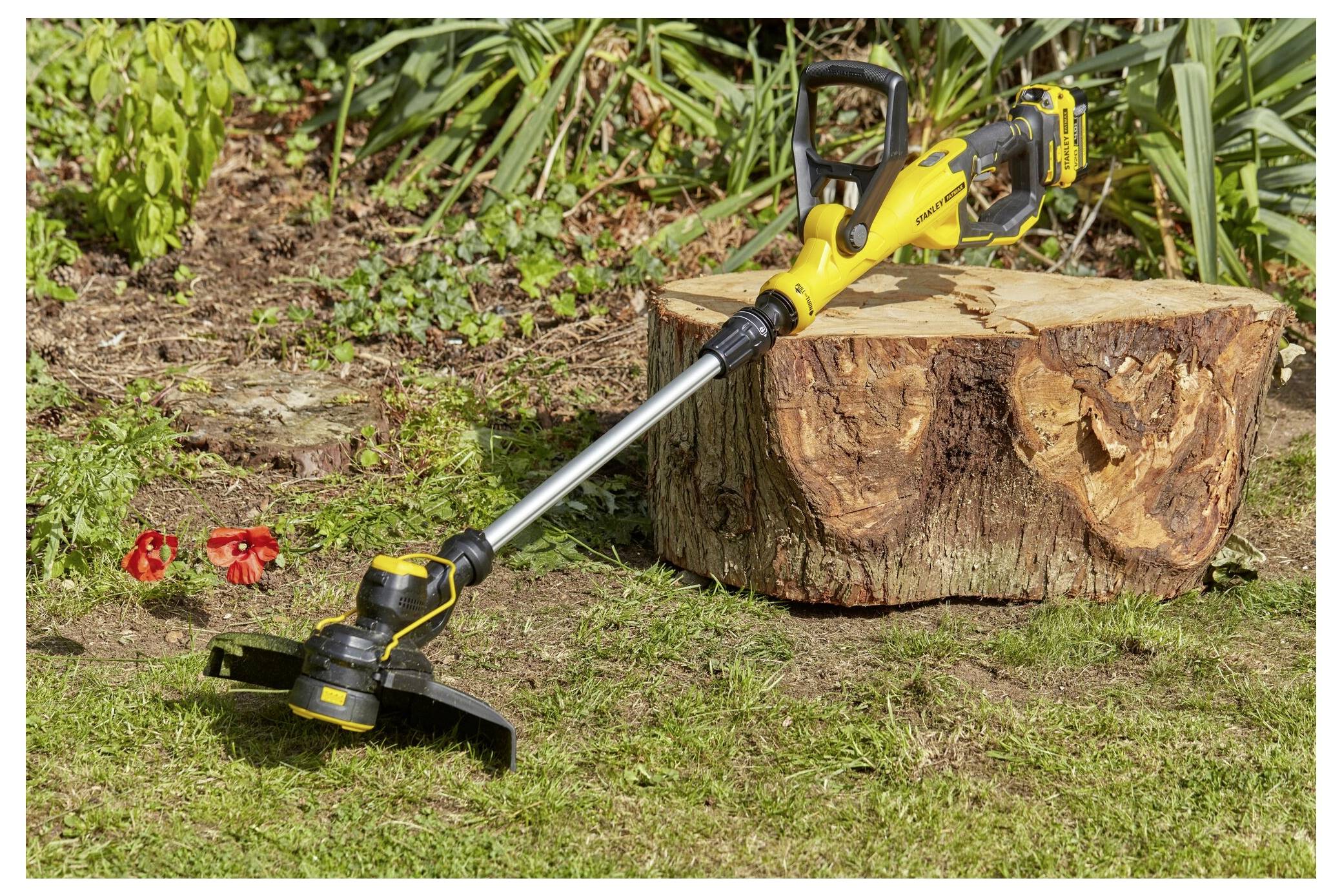 Ein gelb-schwarzer kabelloses Rasentrimmer ruht auf dem Gras neben einem großen Baumstumpf, mit grünen Pflanzen und roten Blumen im Hintergrund.