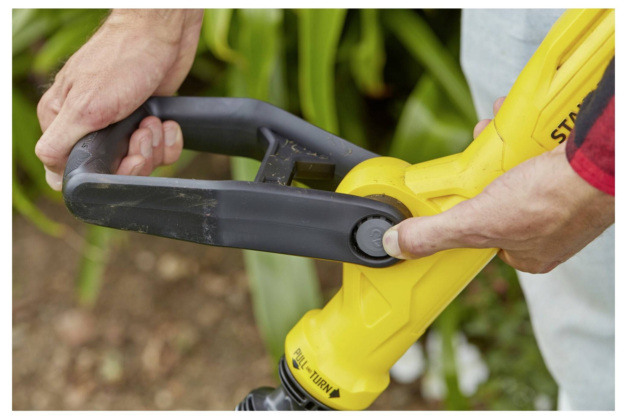 Hände, die einen gelben Fadentrimmer mit Fokus auf den Griff und die Kontrolltaste einstellen, was auf das Einrichten oder Bedienen im Garten hindeutet.