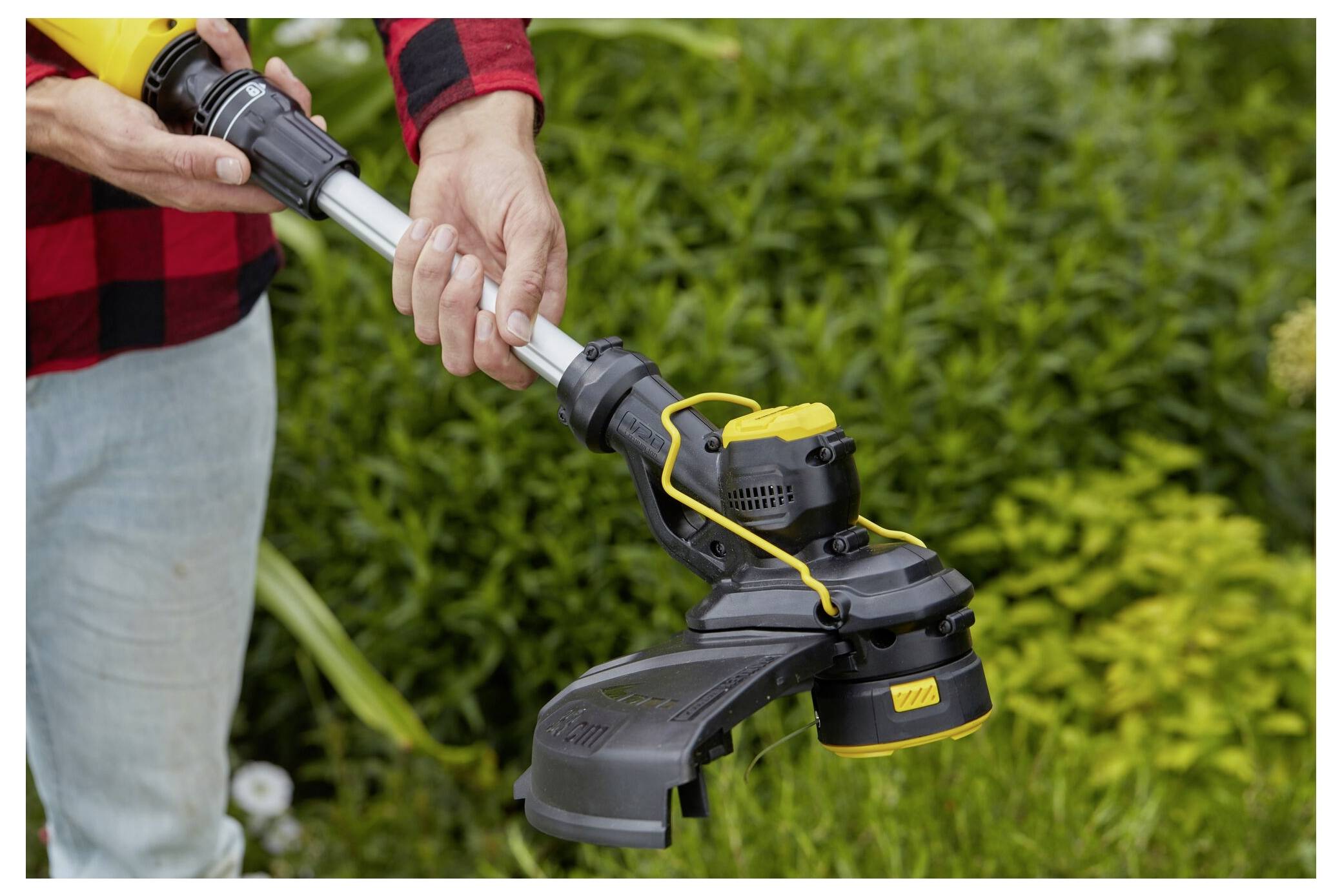 Eine Person in einem rot-schwarzen karierten Hemd hält einen schwarz-gelben elektrischen Rasentrimmer und steht vor einem üppigen grünen Garten.