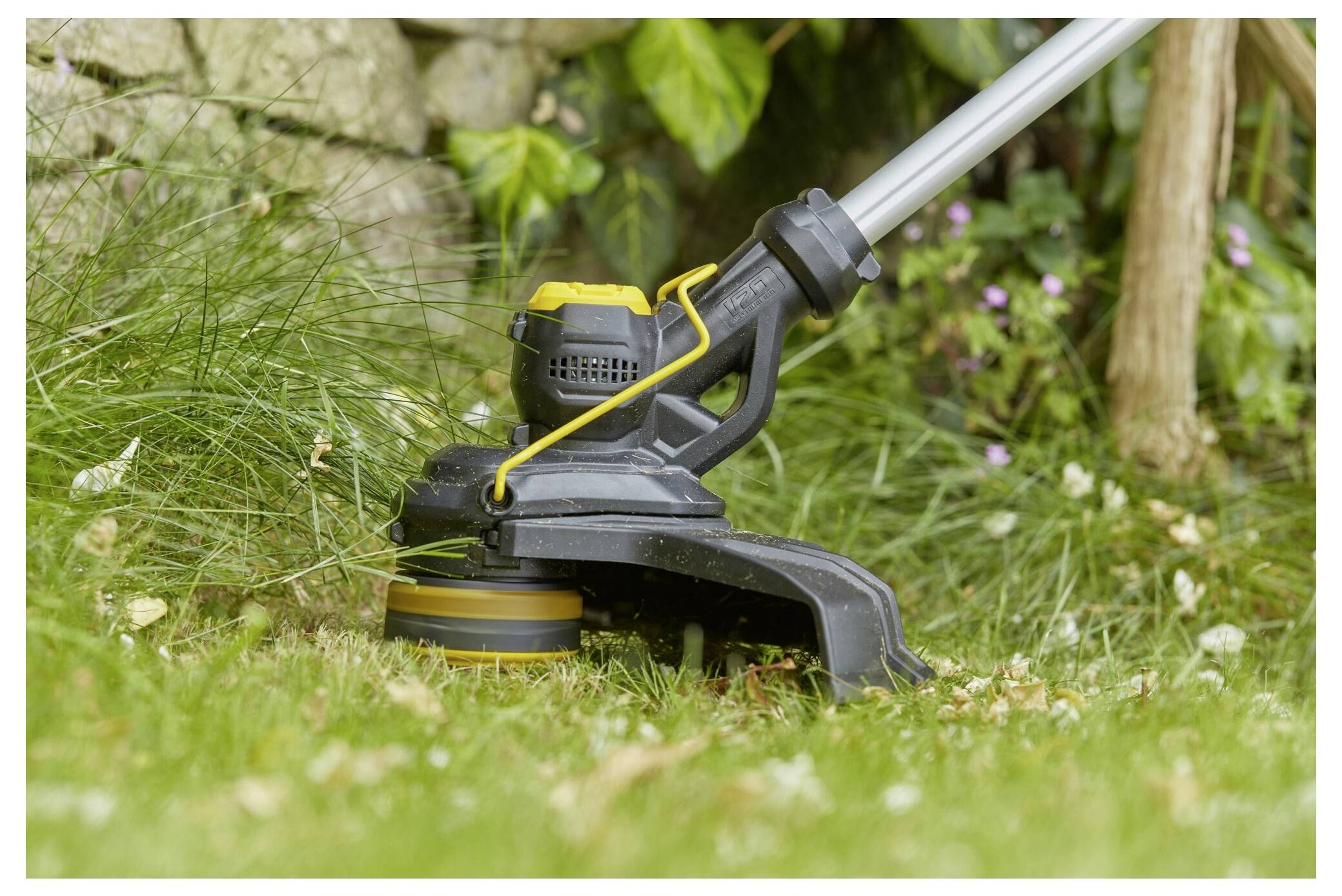 Ein Rasentrimmer schneidet eine Grasfläche in der Nähe einer Steinmauer und Pflanzen und zeigt dessen Verwendung bei der Gartenpflege.