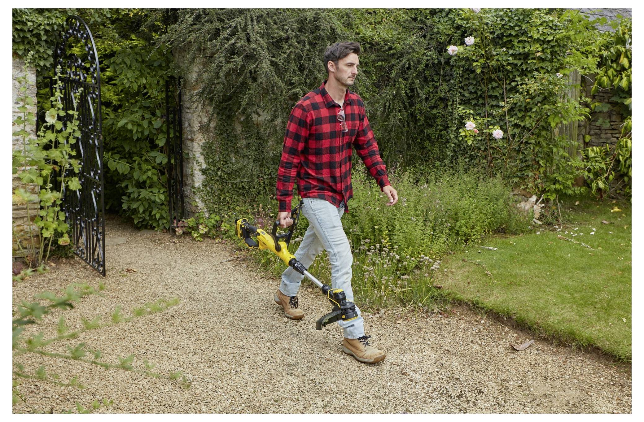 Eine Person geht auf einem Kieselweg und hält eine gelbe Heckenschere, umgeben von grünen Büschen und einem Metallbogen in einer Gartenlandschaft.