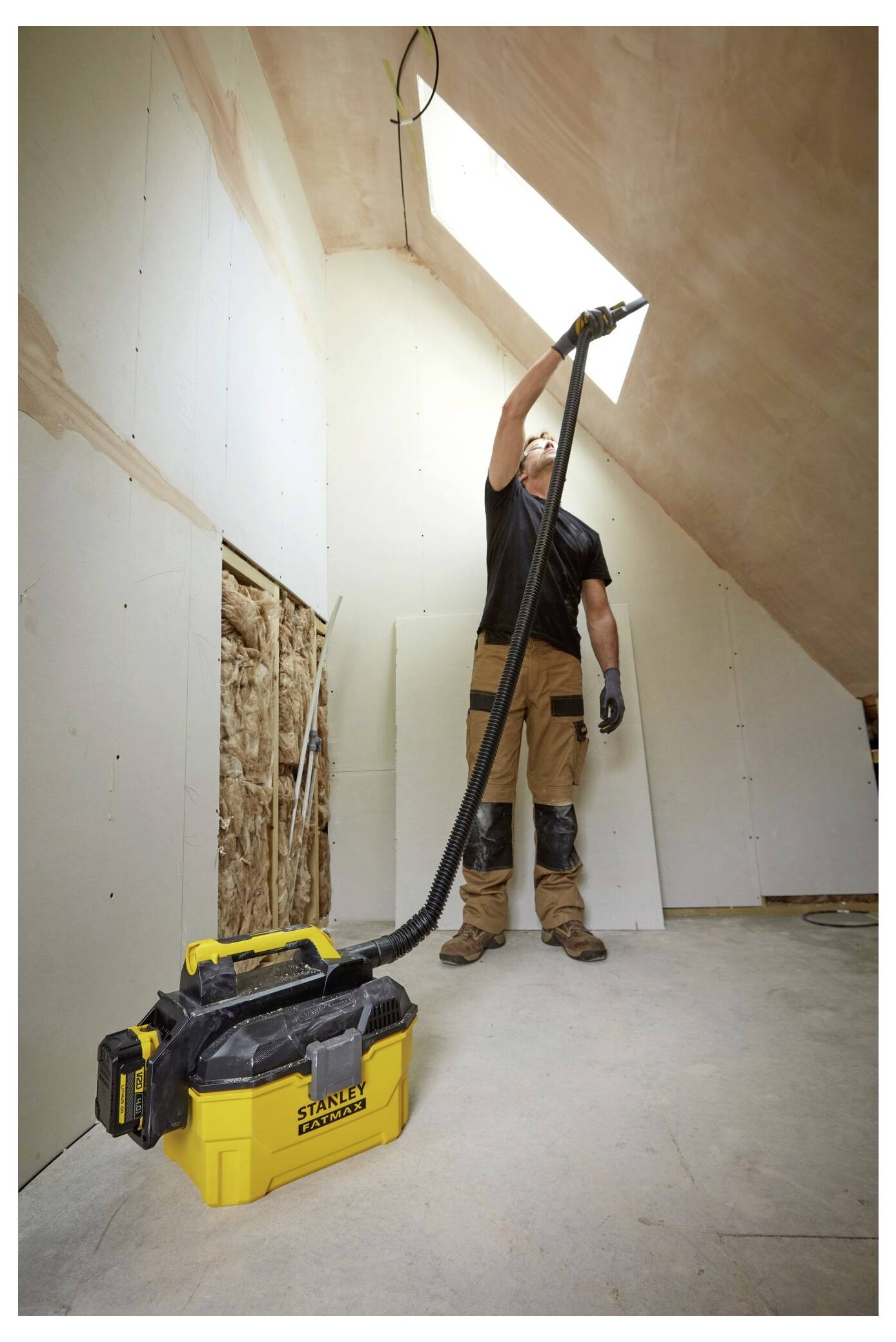 Eine Person, die Trockenbau in einem Zimmer mit schrägen Decken installiert, verwendet einen Industriestaubsauger, um Staub und Schutt zu beseitigen, mit natürlichem Licht von einem Dachfenster.