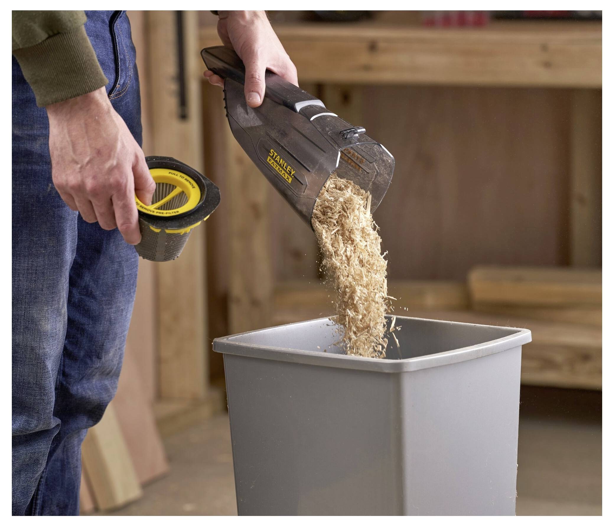 Eine Person leert Sägespäne aus einem Staubsaugerfiler in einen grauen Mülleimer in einer Werkstatt.
