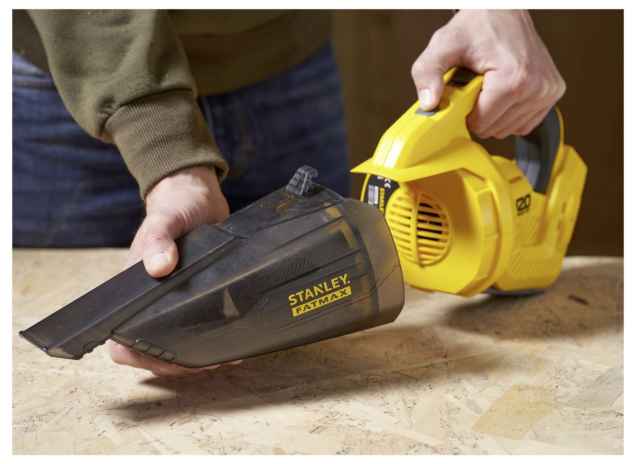 Eine Person entfernt den Staubbehälter aus einem gelben Stanley Fatmax tragbaren Staubsauger auf einer Holzoberfläche.