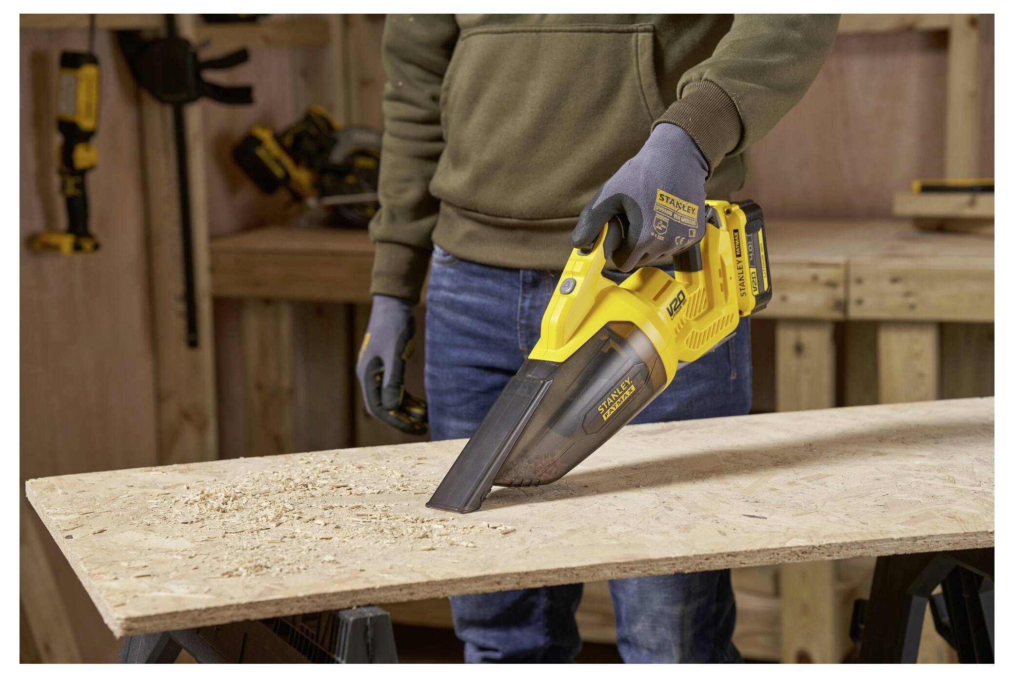 Eine Person benutzt einen gelben tragbaren Staubsauger, um Holzspäne auf einer Werkbank in einer Werkstatt zu reinigen. Im Hintergrund hängen Werkzeuge an der Wand.