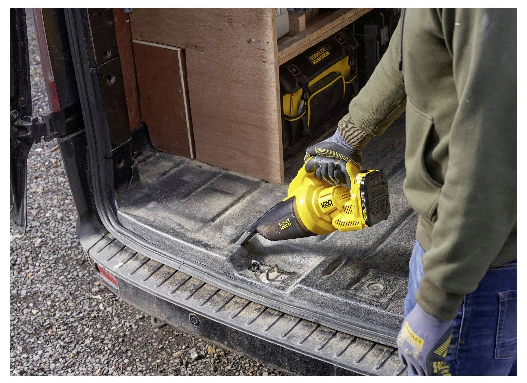Eine Person mit Handschuhen benutzt einen gelben schnurlosen Staubsauger, um den Kofferraum eines Fahrzeugs zu reinigen, während Werkzeuge im Hintergrund sichtbar sind.