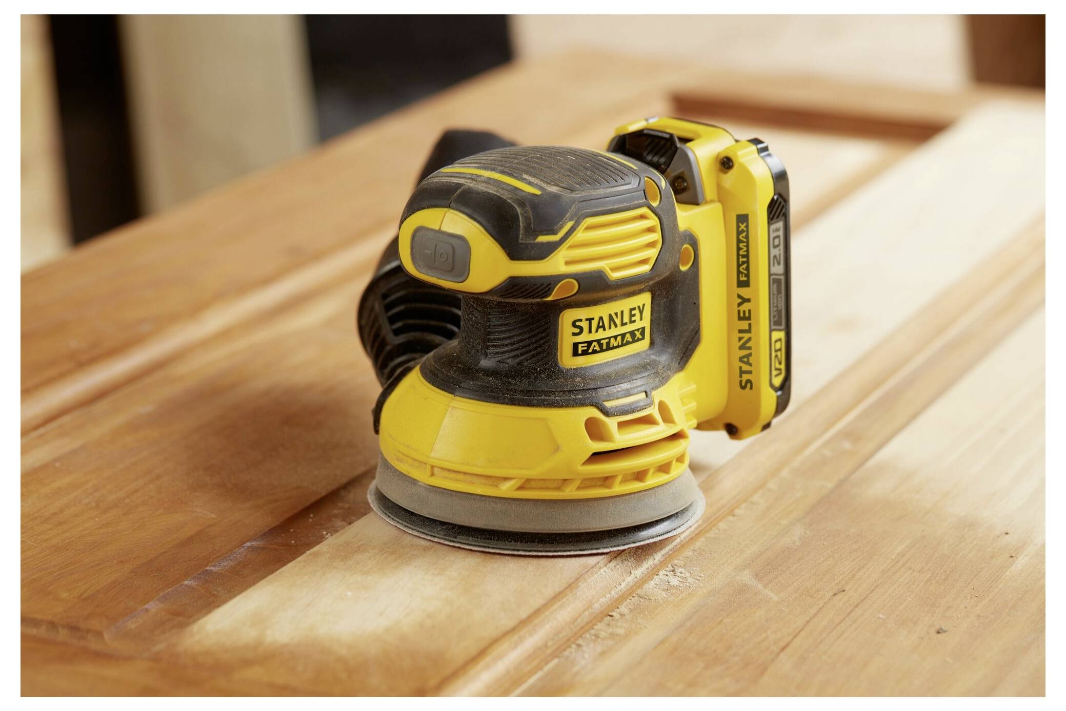 Ein gelb-schwarzer Stanley FatMax kabelloses Elektroschliff-Exzenterschleifer sitzt auf einer Holzoberfläche und hebt sein ergonomisches Design und die Batterieplatzierung hervor.