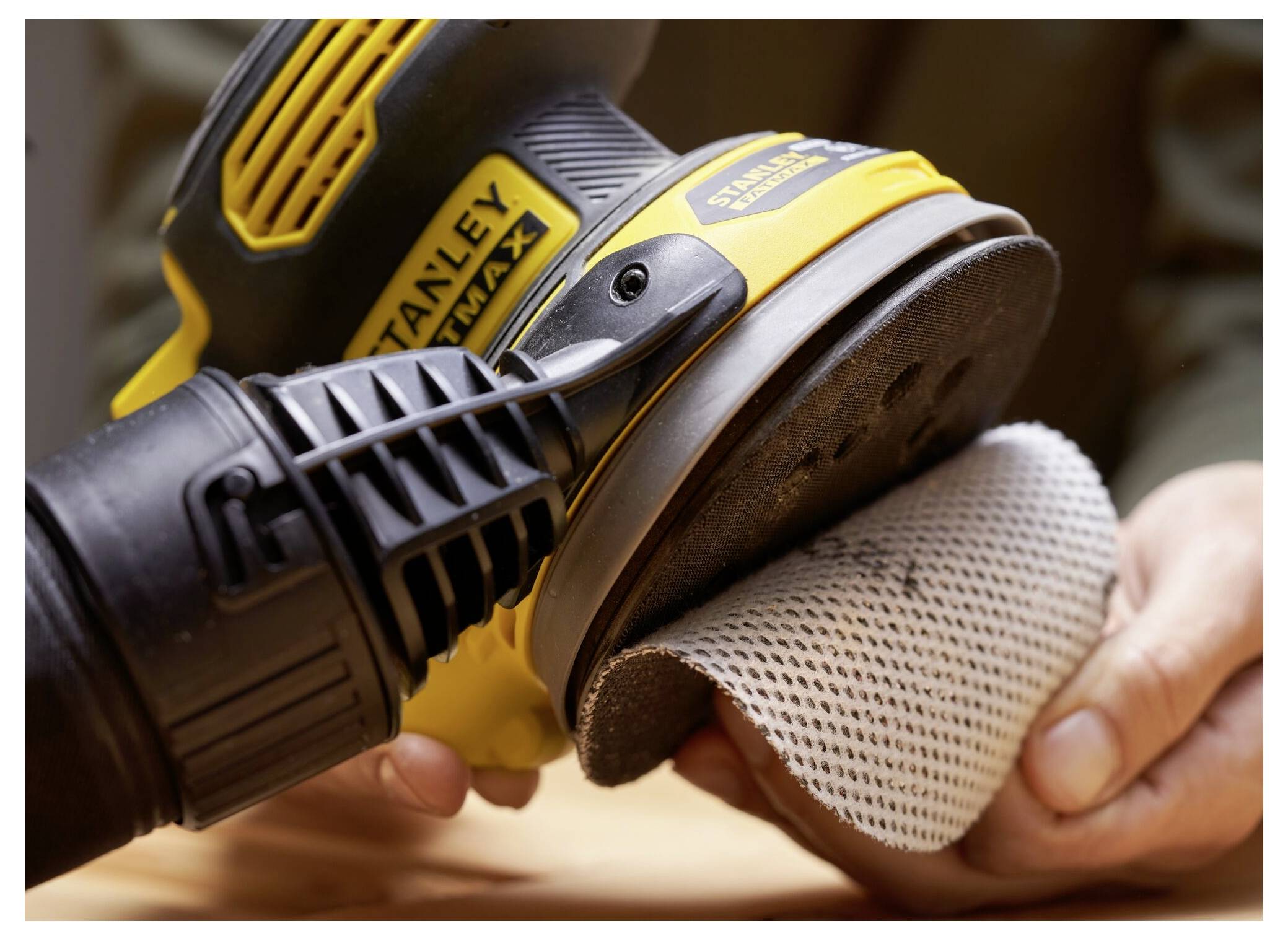 Eine Person verwendet einen gelb-schwarzen Elektroschleiffer, um ein Stück Holz zu glätten, und drückt das Werkzeug fest gegen das Schleifpapier.