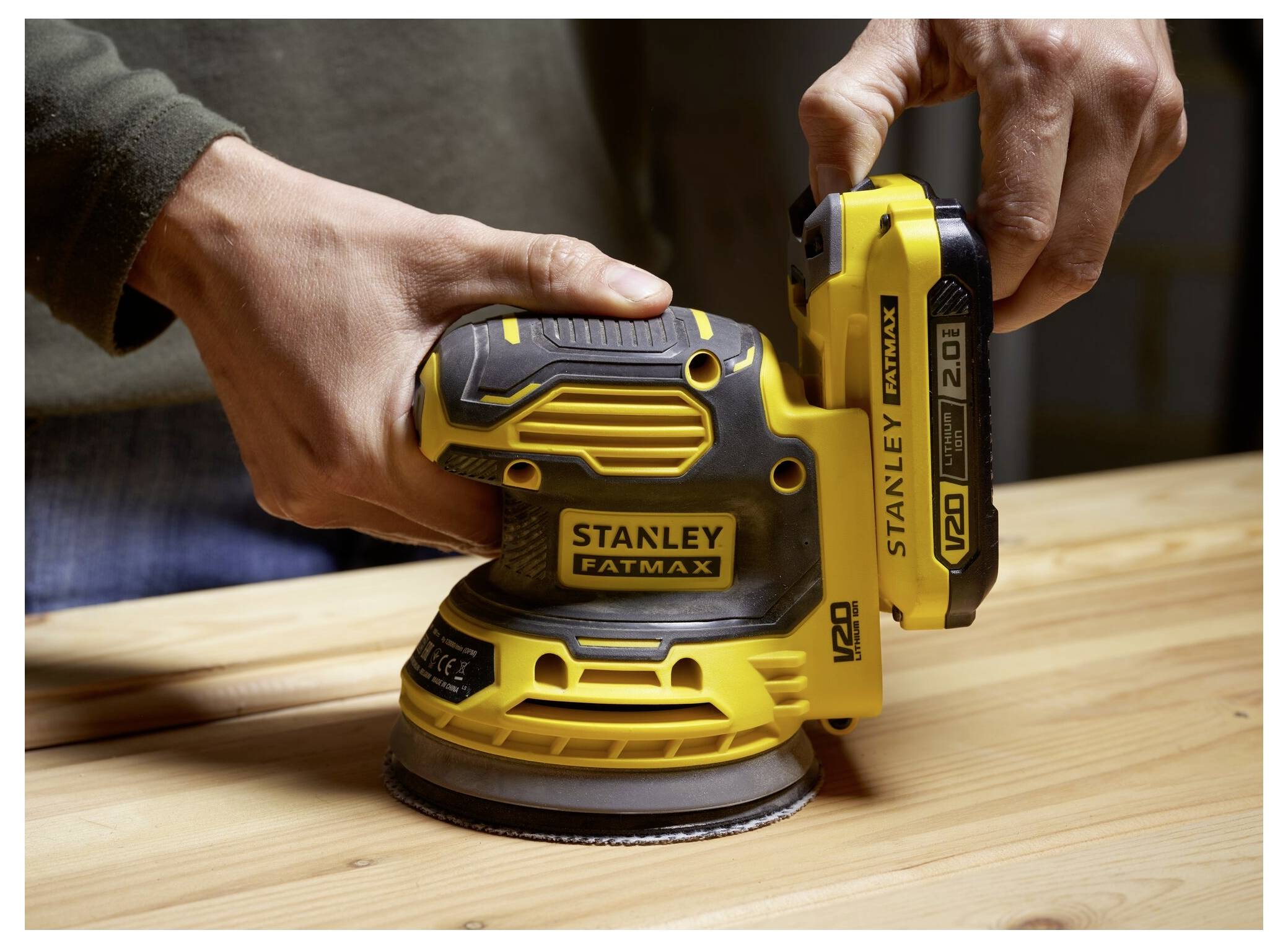Eine Person, die einen gelb-schwarzen Stanley Fatmax Schleifer auf einer Holzoberfläche benutzt, wobei die Werkzeugdetails und die Holzbearbeitung betont werden.