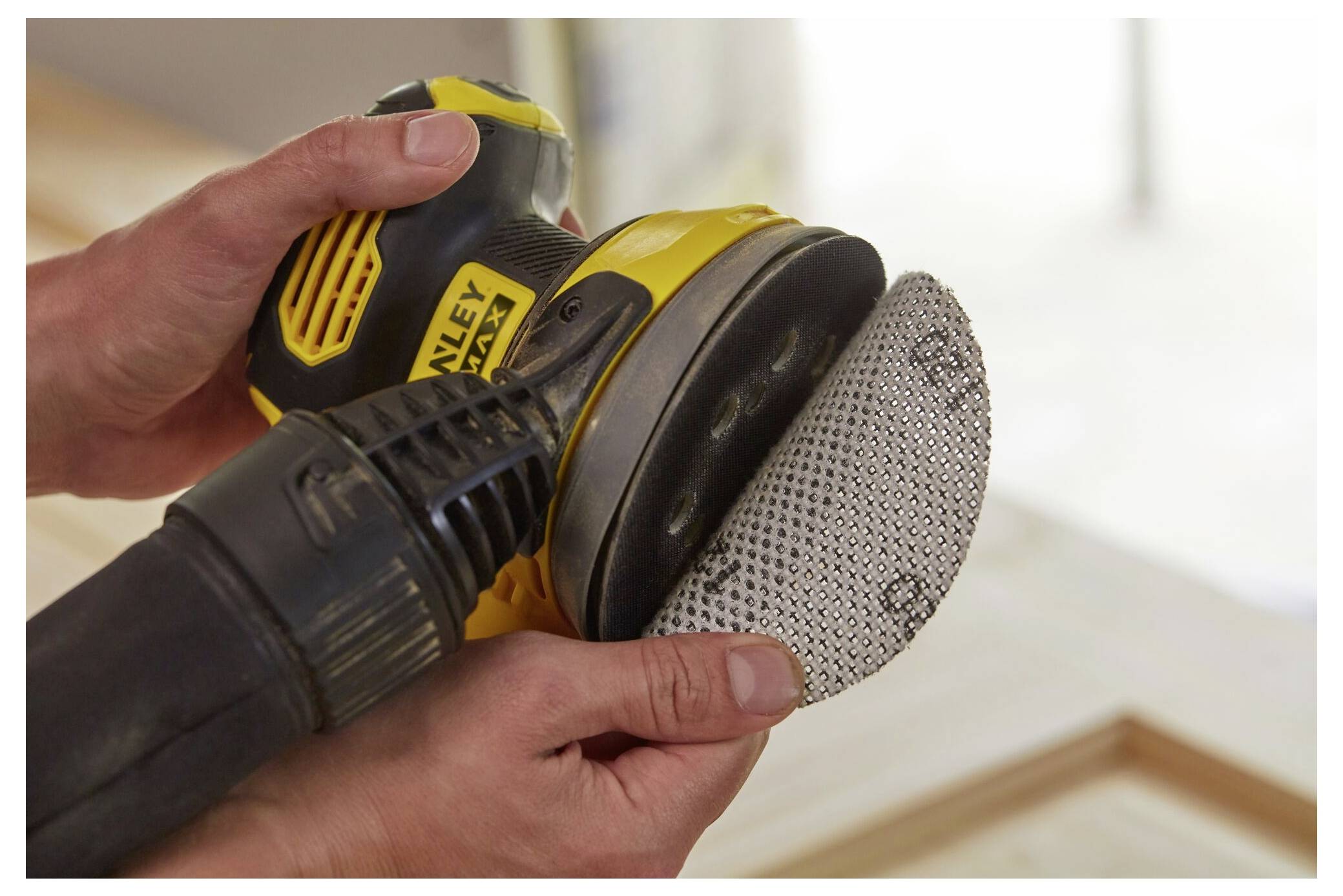 Hände halten einen gelb-schwarzen Elektroschleiffer mit einer kreisförmigen Schleifplatte, die gerade befestigt wird, was eine Vorbereitung zum Holzbearbeiten andeutet.