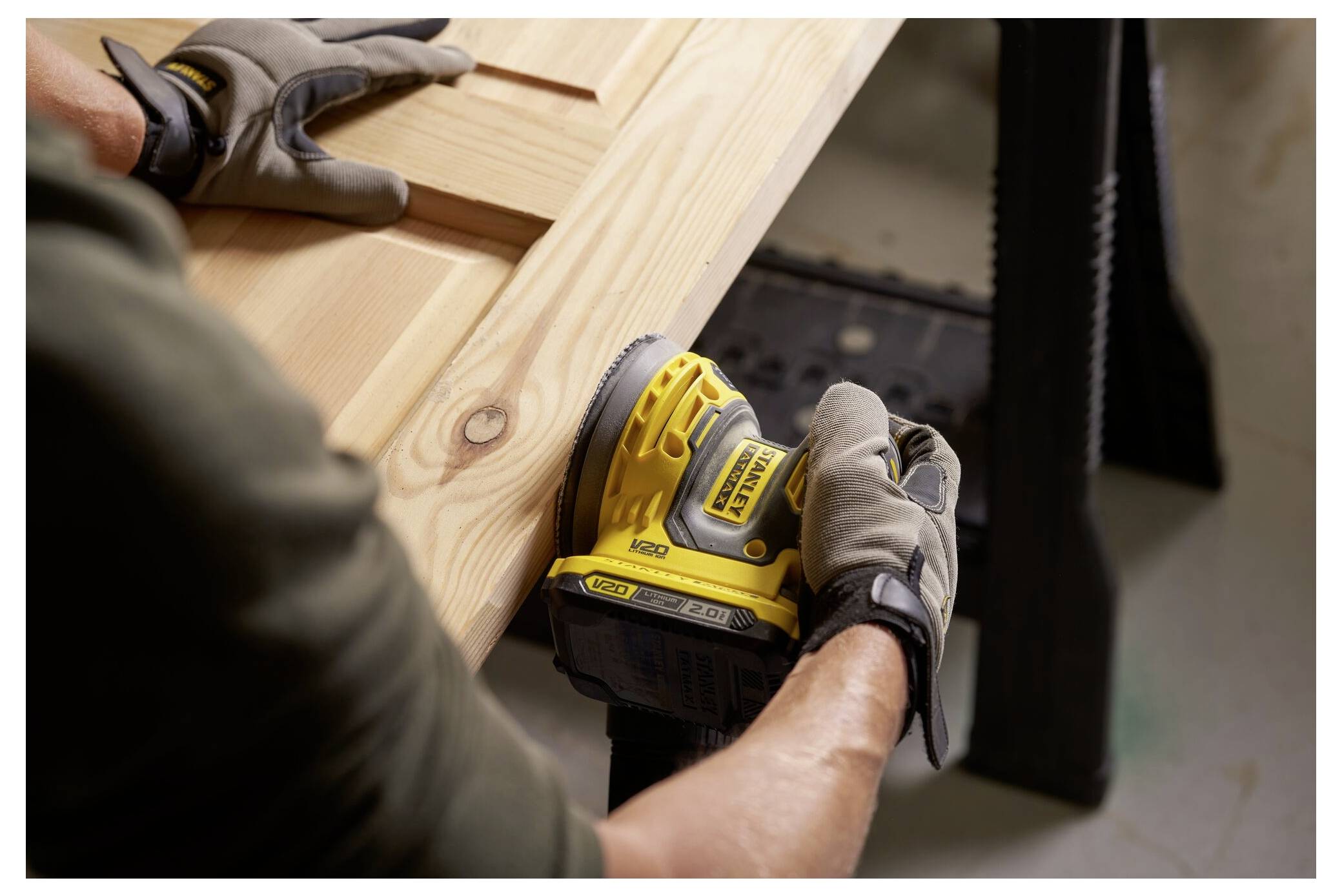 Person, der einen gelben Elektroschleifera verwendet, um ein Holzbrett auf einem Tisch zu glätten, trägt Handschuhe zum Schutz.