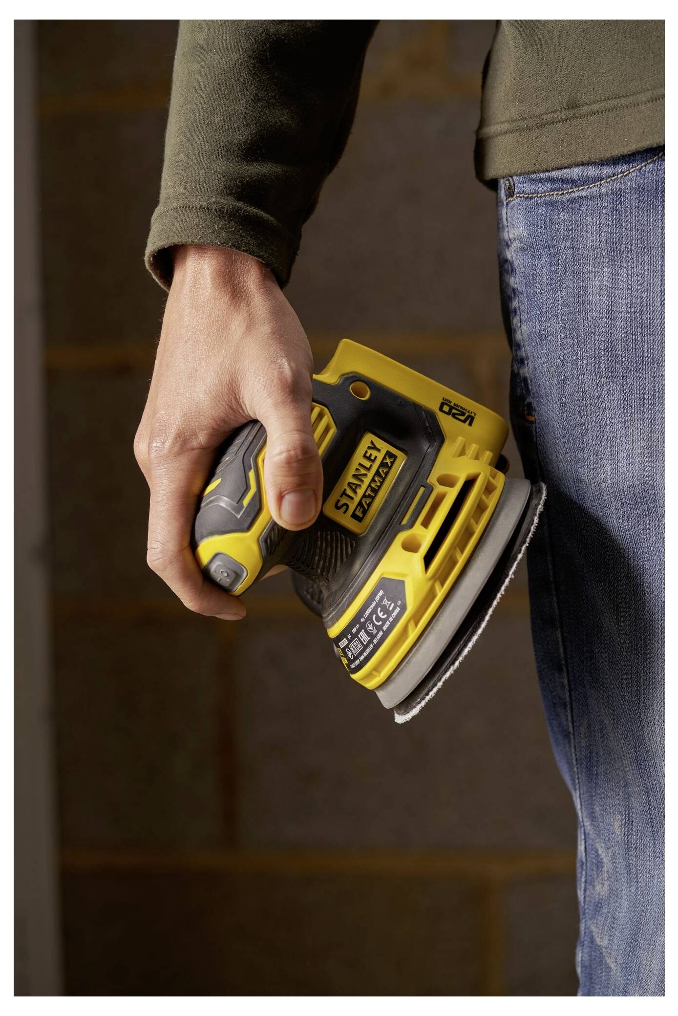 Eine Person hält einen gelb-schwarzen Elektroschleiffer gegen ihr Bein, trägt Jeans und ein dunkles Langarmshirt.