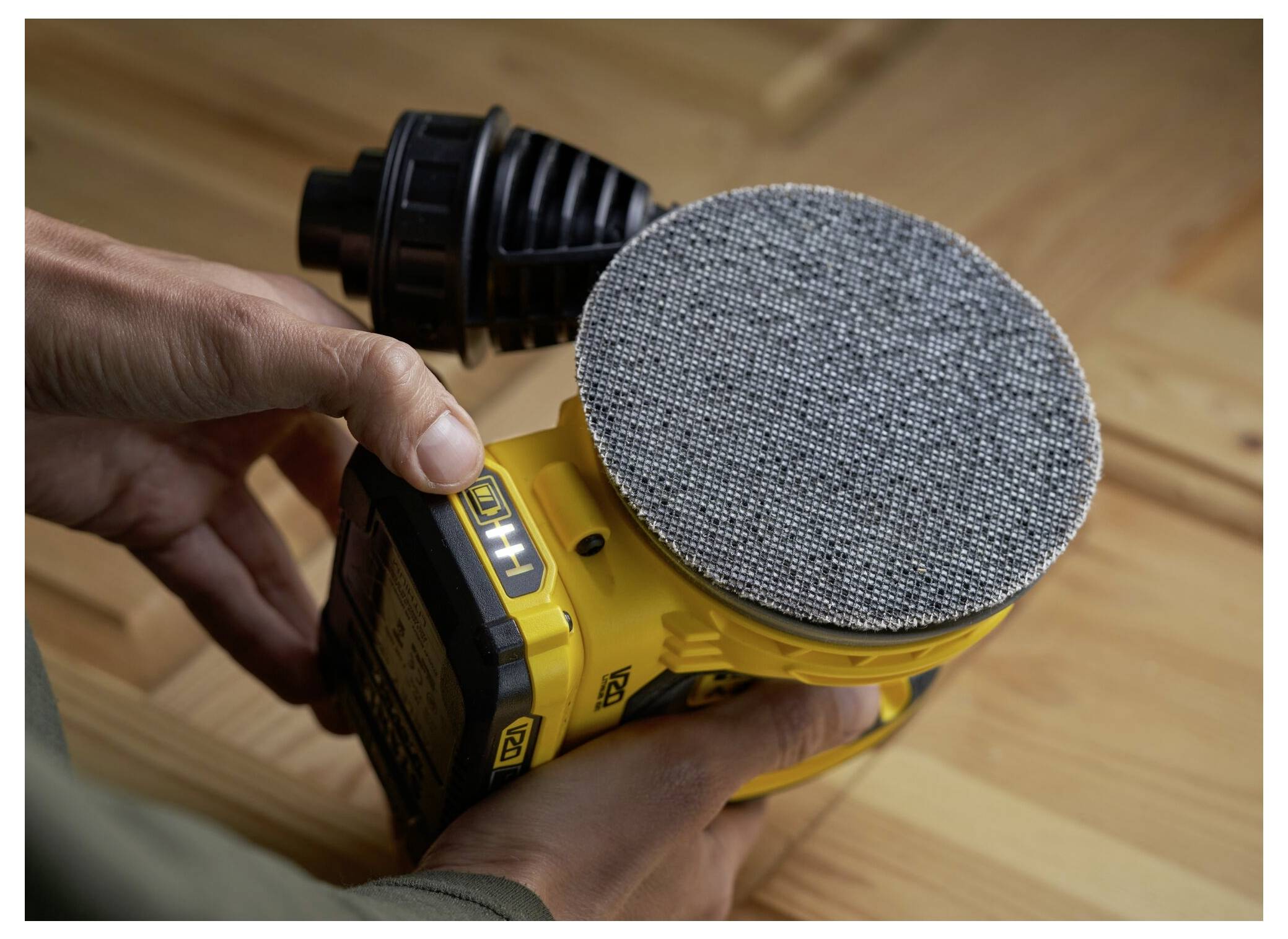 Eine Person hält einen gelben Elektroschleiffer gegen eine Holzoberfläche und zeigt das strukturierte, kreisförmige Schleifpad des Schleifers.