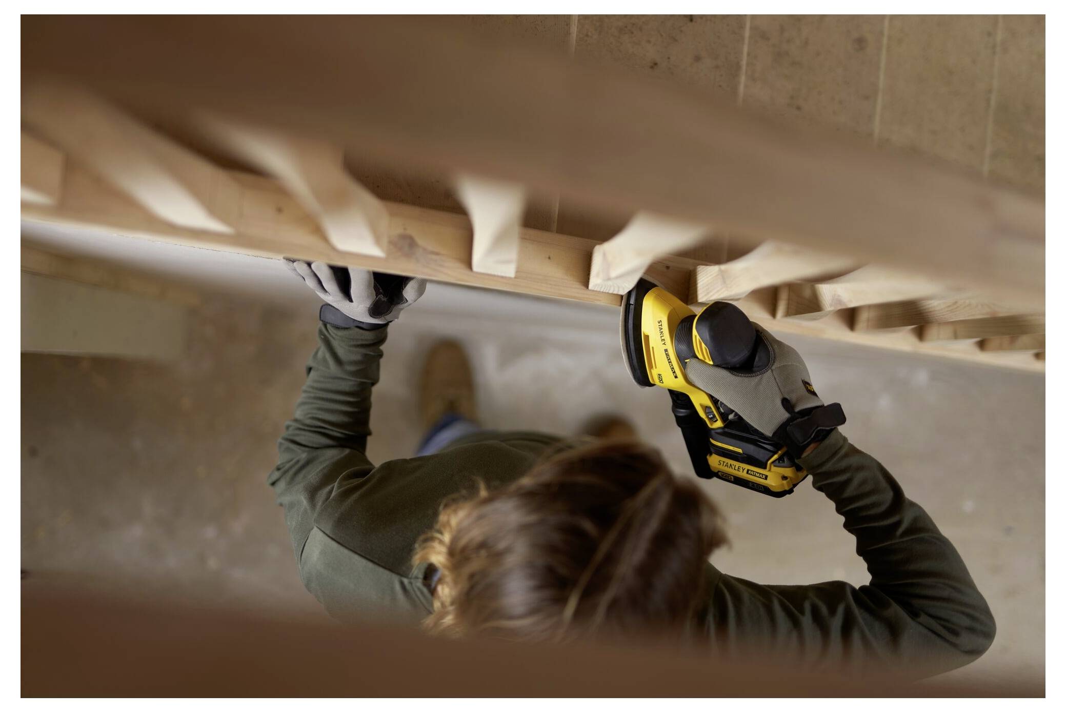 Eine Person, die ein gelbes Elektrowerkzeug auf Holztreppen benutzt, von oben betrachtet. Sie trägt Handschuhe und scheint das Holz zu glätten oder zu schleifen.