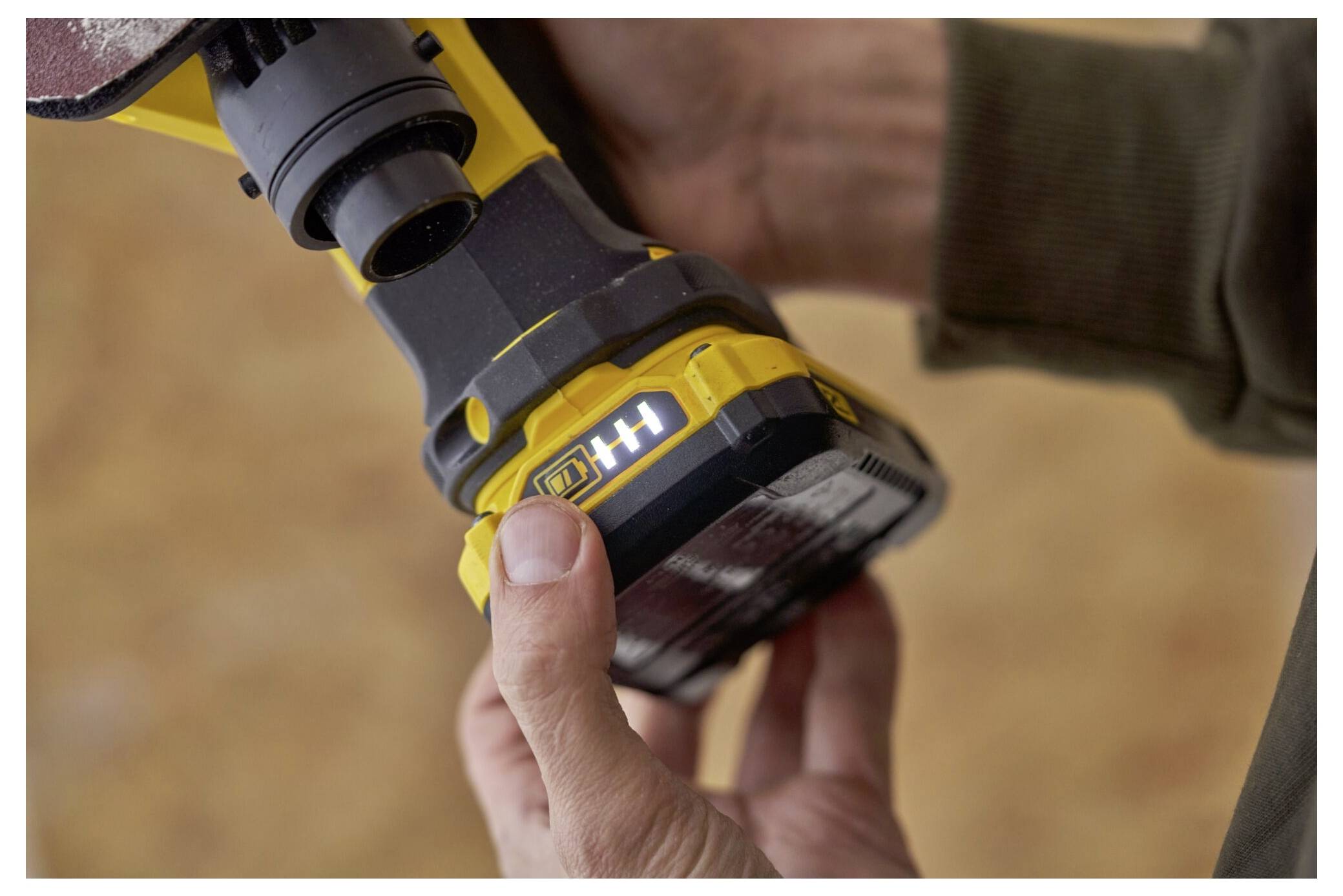 Eine Person hält ein Elektrowerkzeug und untersucht seine Batterieanzeige, die eine vollständige Ladung auf einem gelben, tragbaren Gerät anzeigt.